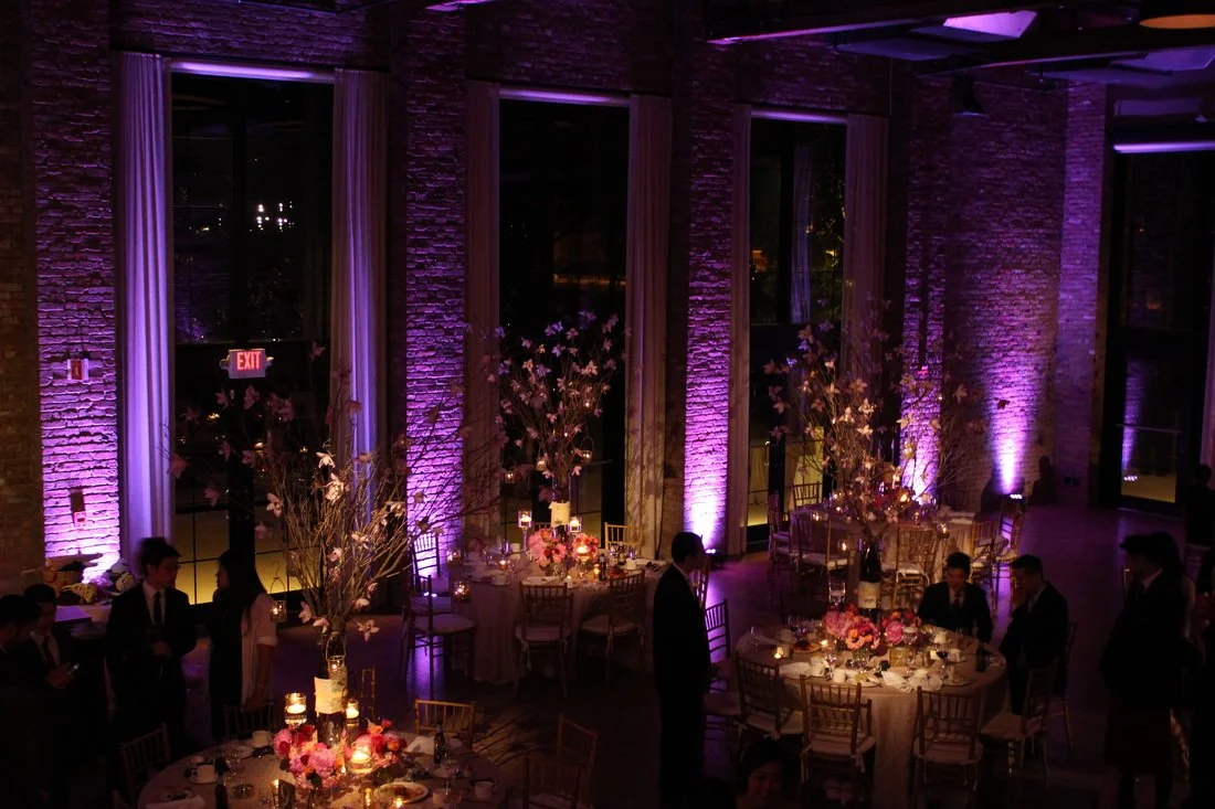 Elegant event venue with purple uplighting on brick walls, tall floral centerpieces, and decorated round tables with chairs, where guests are dressed formally.