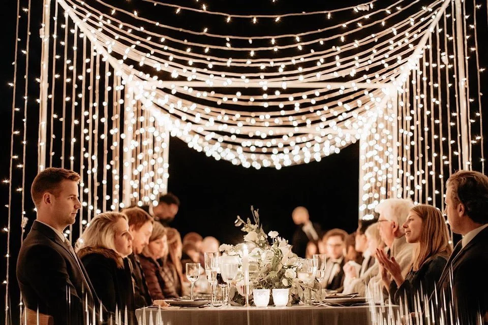 A wedding reception with a long table decorated with floral arrangements and candles, under a canopy of hanging string lights, with guests dressed in formal attire.