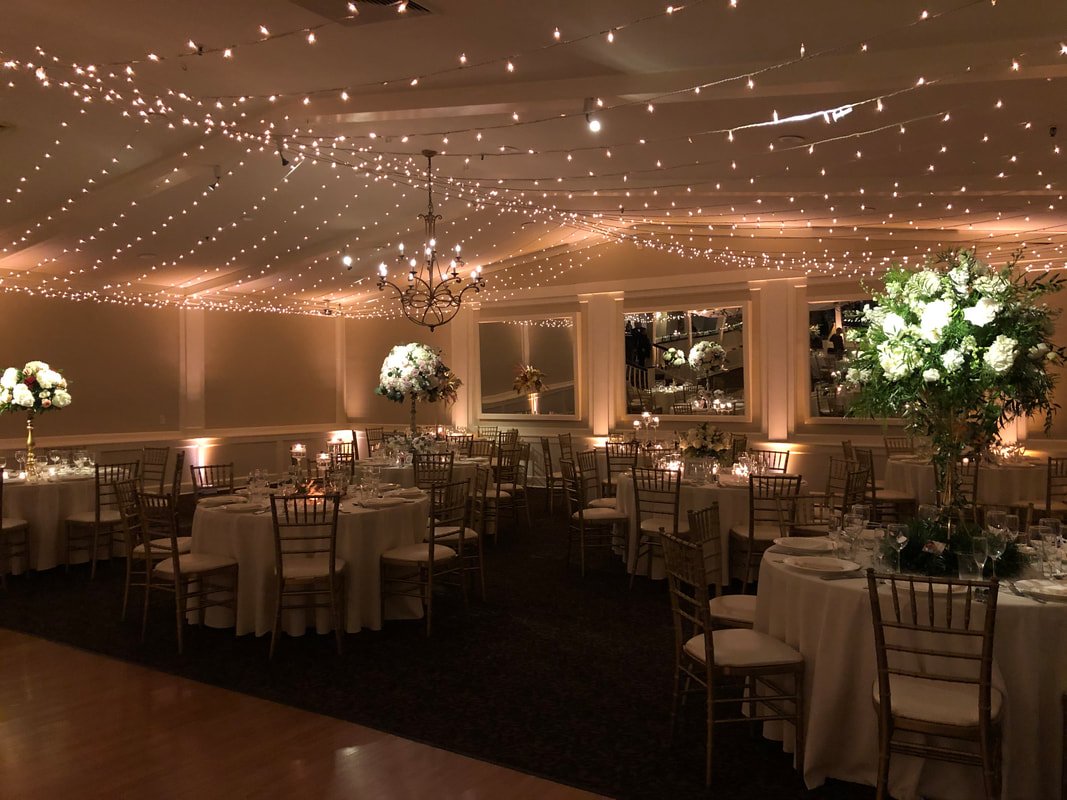 Elegant banquet hall decorated for a formal event with round tables, white tablecloths, floral centerpieces, string lights on the ceiling, and warm ambient lighting.