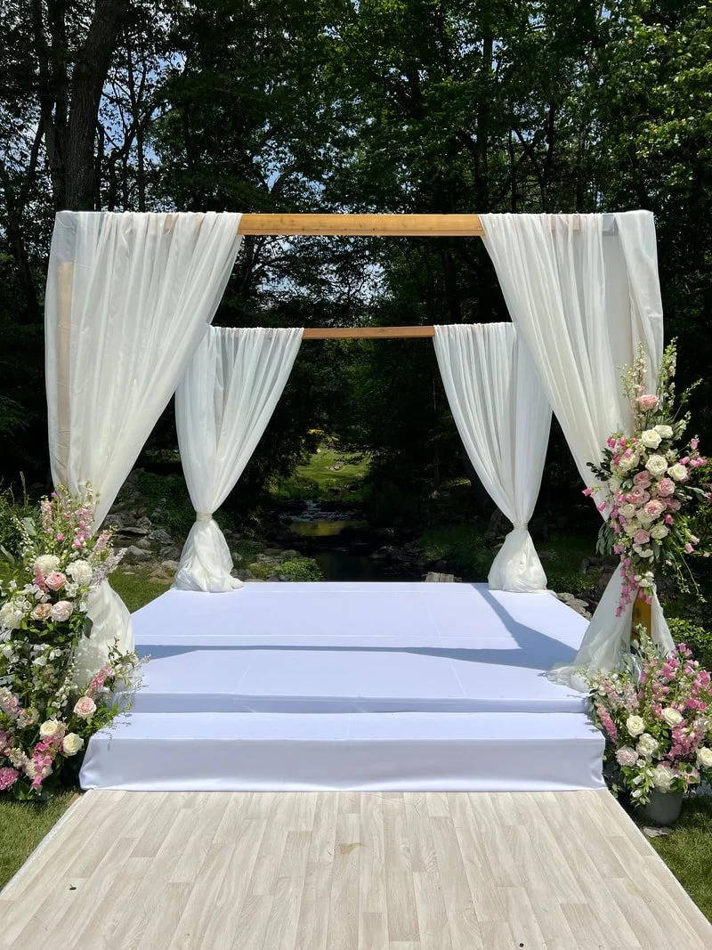 A wedding altar decorated with white curtains and floral arrangements on both sides, set outdoors with a natural forest background.