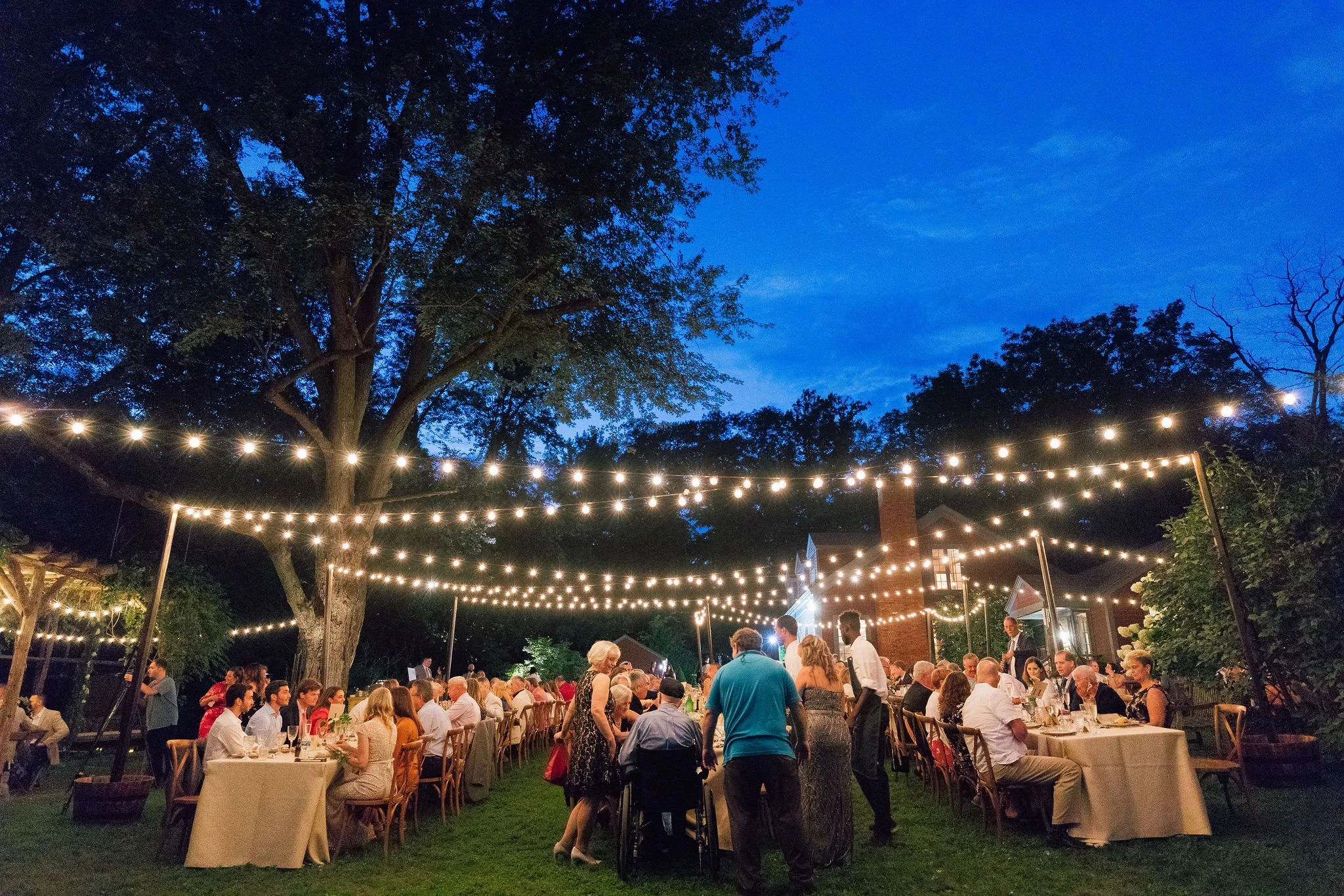 Outdoor evening gathering under string lights with people seated at tables and socializing, surrounded by large trees and a house in the background.