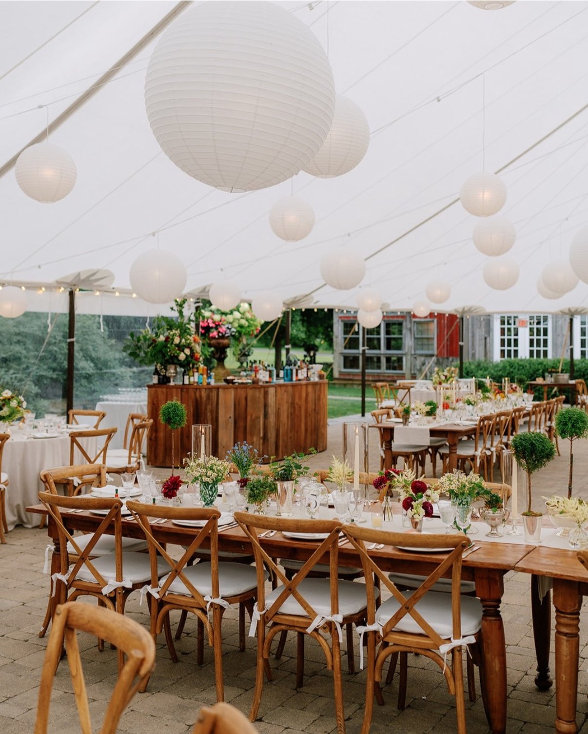 Decorated wedding reception under a tent with round paper lanterns, long wooden tables with floral centerpieces, and wooden chairs with white cushions.