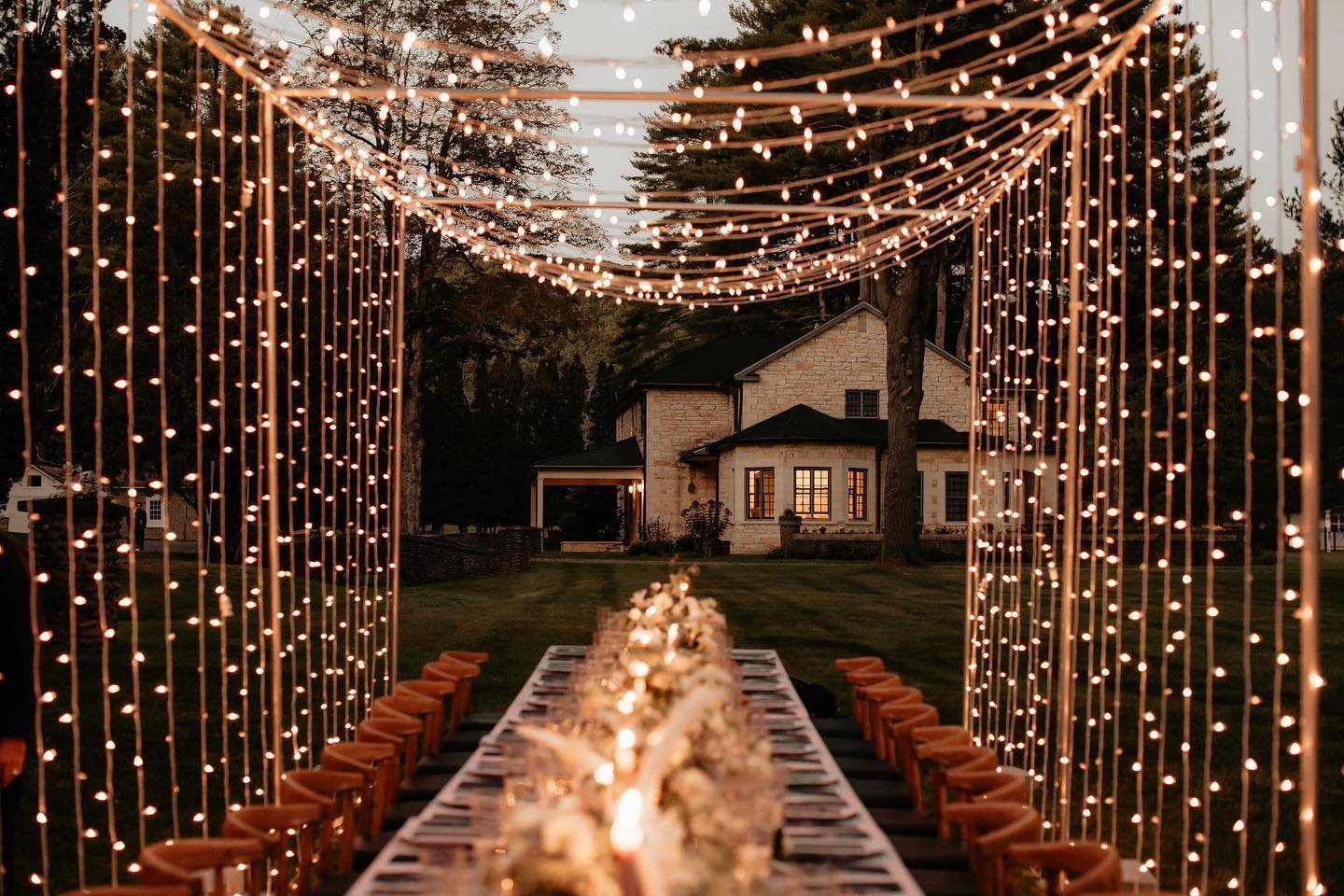 An elegant outdoor dinner setup under a canopy of hanging string lights at dusk, with a long table decorated with flowers, leading towards a large house in the background.