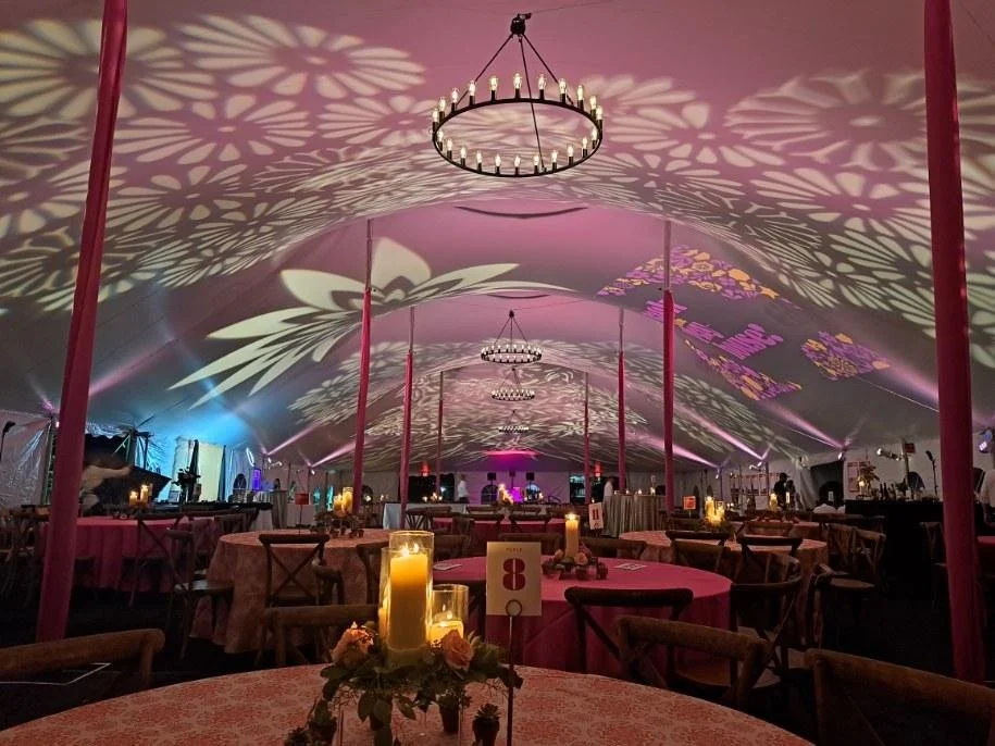 Interior of a decorated event tent with pink and purple lighting, circular tables with candles and flower centerpieces, and chandeliers casting floral shadow patterns on the ceiling.