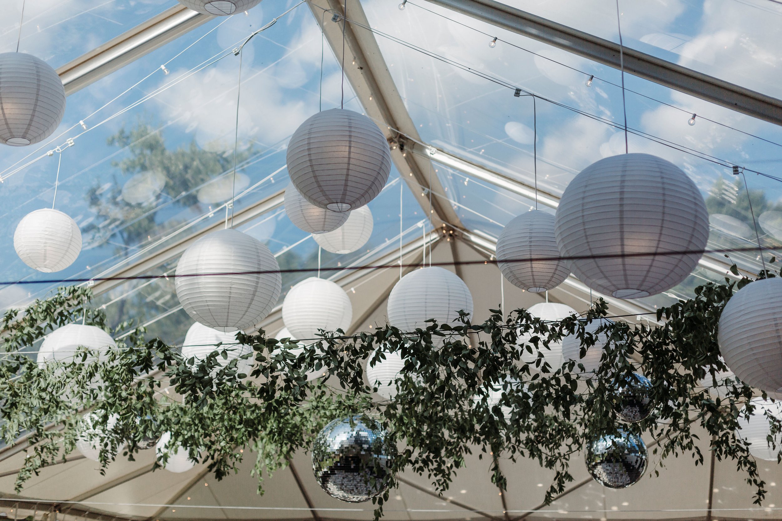 Decorative white paper lanterns and silver disco balls hanging from strings and greenery inside a transparent tent with a view of a blue sky and clouds outside.