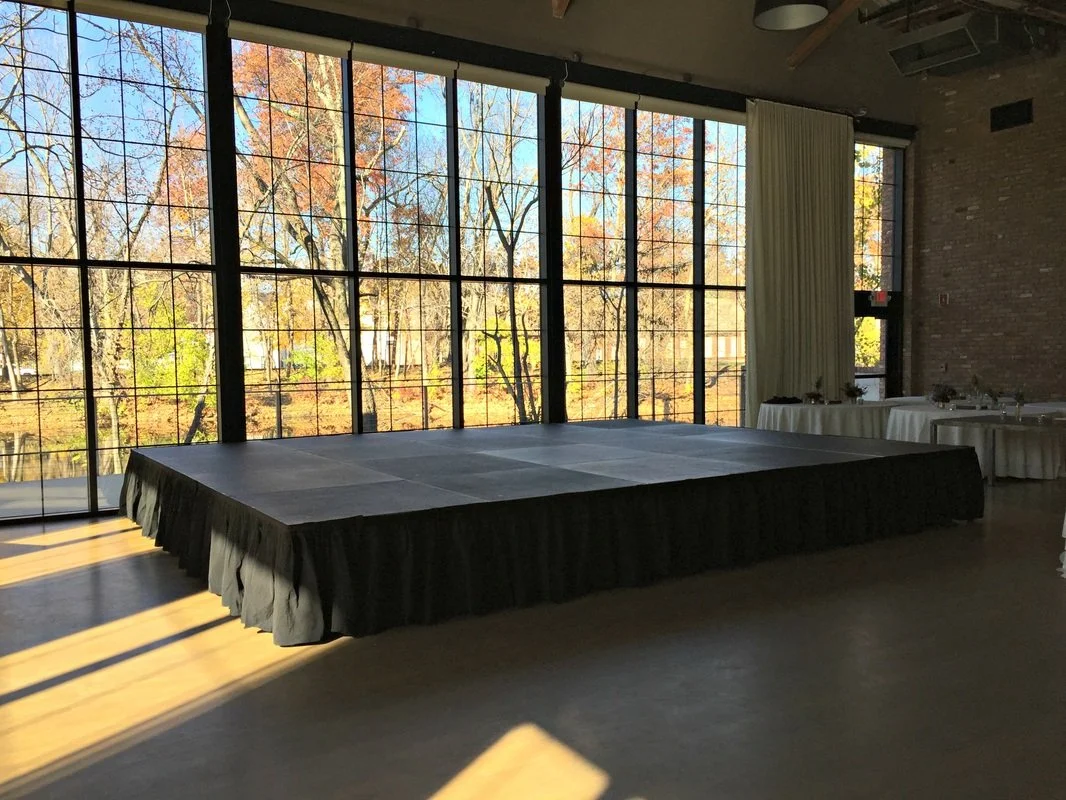 Empty event space with a large, black-draped stage and floor-to-ceiling windows showing trees with autumn foliage.