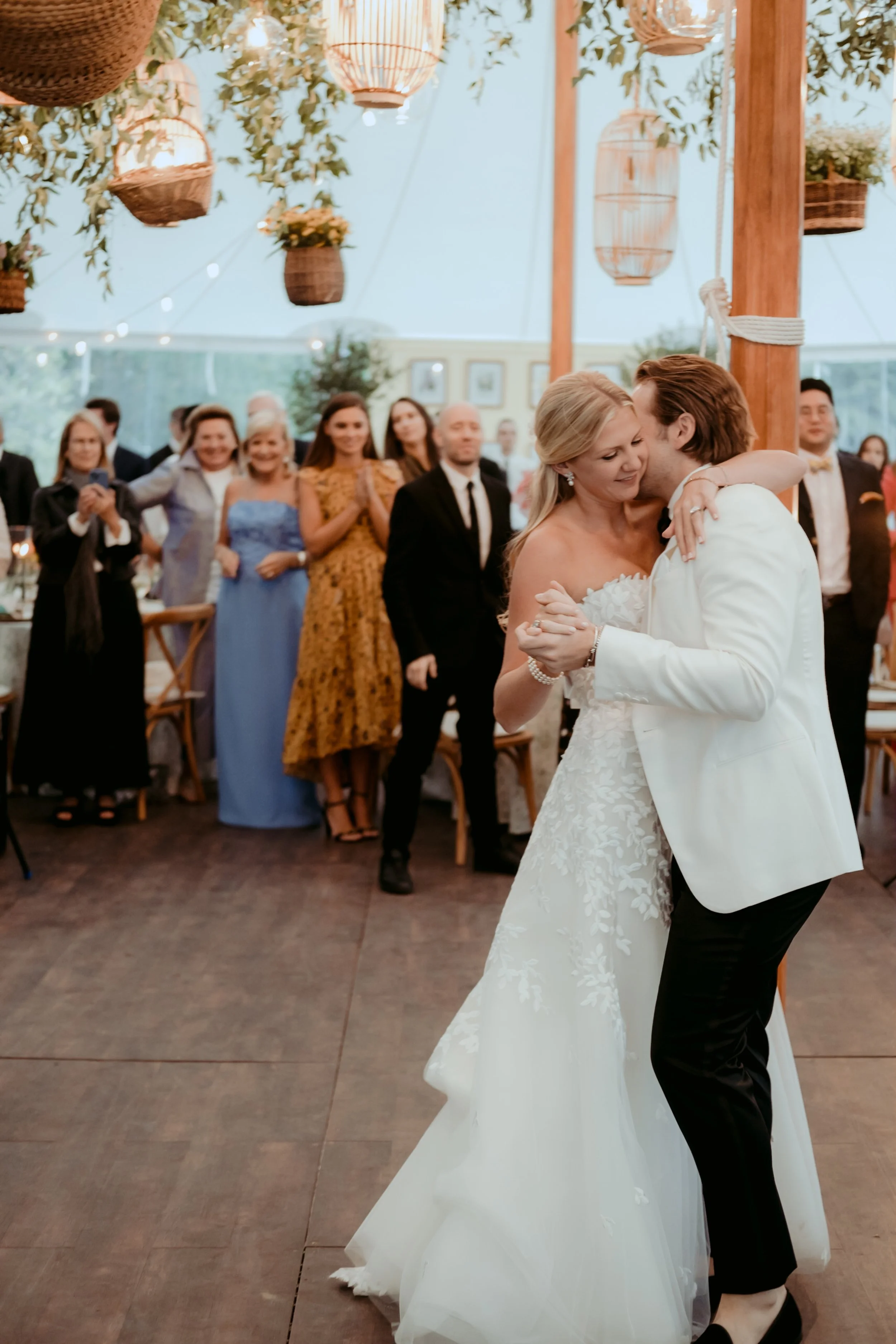 A bride and groom dance closely at their wedding reception, with guests clapping and smiling in the background, decorated with hanging lanterns and potted plants.