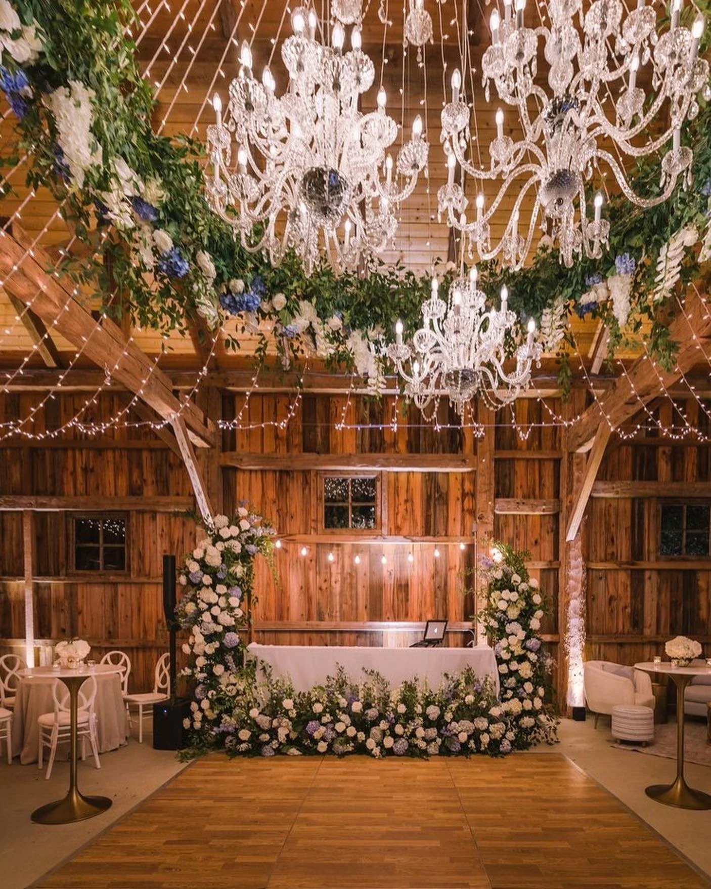 Elegant wedding or event venue with a wooden barn interior, decorated with large crystal chandeliers hanging from the ceiling, string lights, floral arrangements of white and purple flowers, and a decorated table with a laptop in the center.