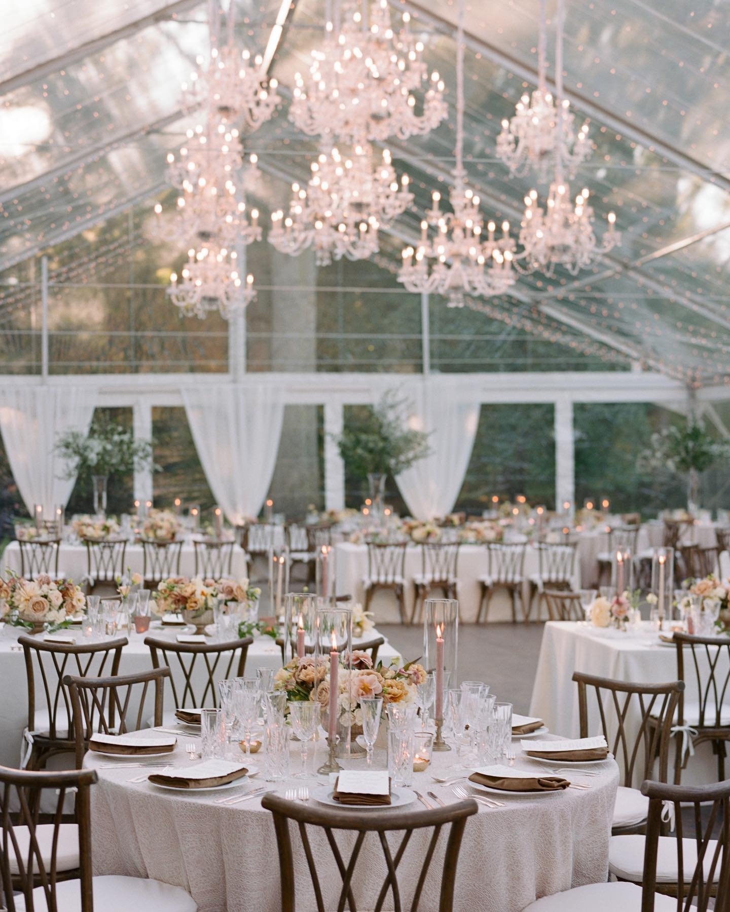 Elegant indoor wedding reception with round and long banquet tables decorated with floral centerpieces, tall candles, and glassware, under a transparent ceiling with chandeliers and string lights.