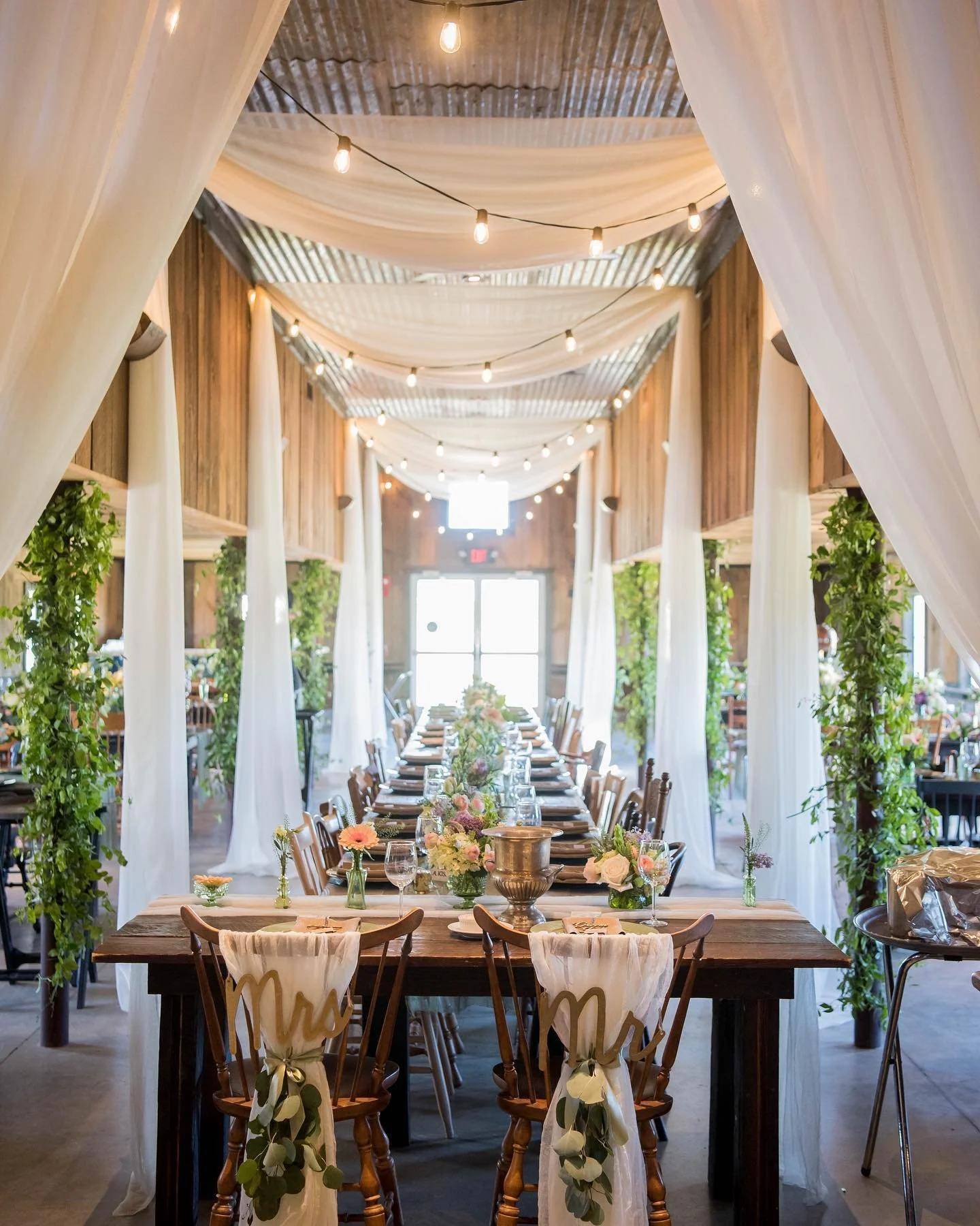 Long banquet table decorated with flowers and candles in a rustic, decorated event space with draped fabric and string lights.