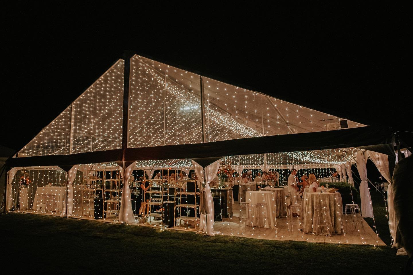Nighttime outdoor wedding reception with string lights inside a large tent with tables, people, and floral arrangements.
