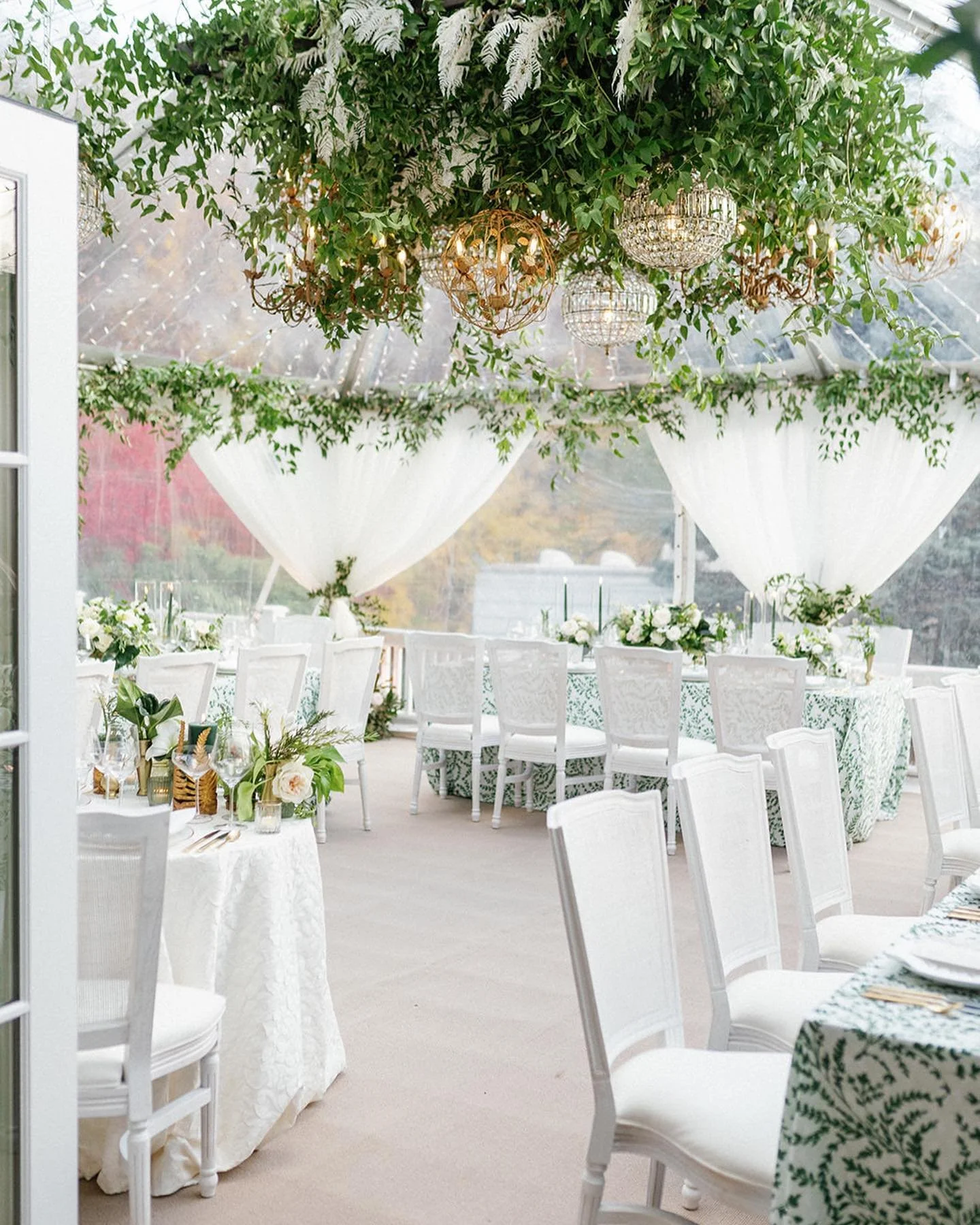 Elegant wedding reception setup inside a glass tent with white chairs, floral arrangements, green foliage, and hanging chandeliers.