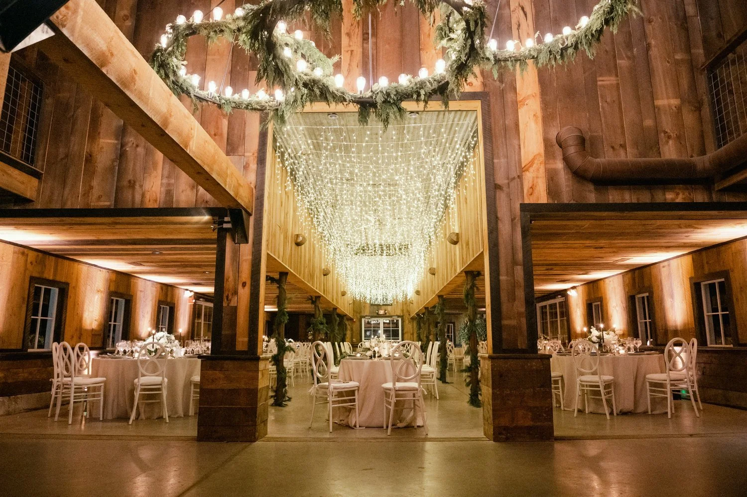 Decorated banquet hall with round tables covered in white tablecloths, elegant chairs, and a high ceiling adorned with strings of warm fairy lights. The room has wooden walls and festive greenery with candles on the tables.