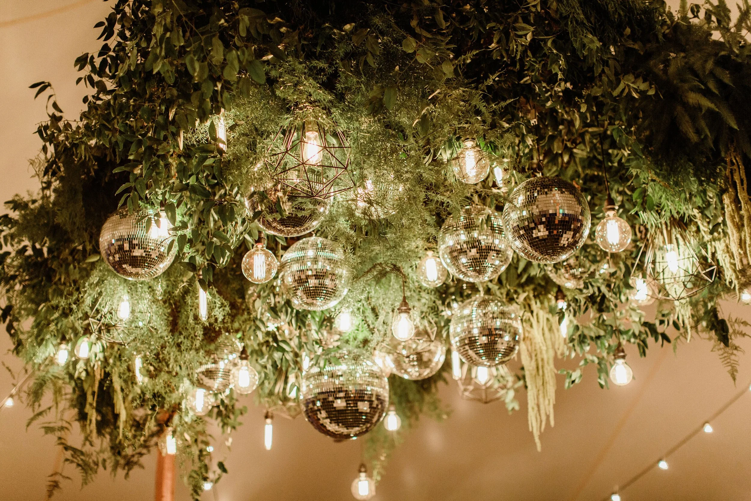 Decorative ceiling installation with hanging disco balls, light bulbs, and greenery