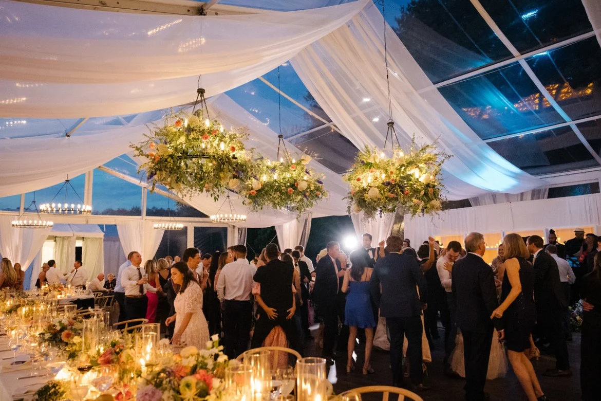 Guests dancing and socializing at a wedding reception inside a decorated tent with hanging floral chandeliers and draped fabric ceiling.