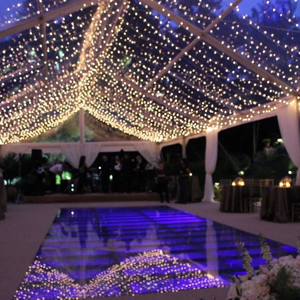 String of hanging string lights under a tent, reflected on a glossy dance floor at night, with tables and people in the background.