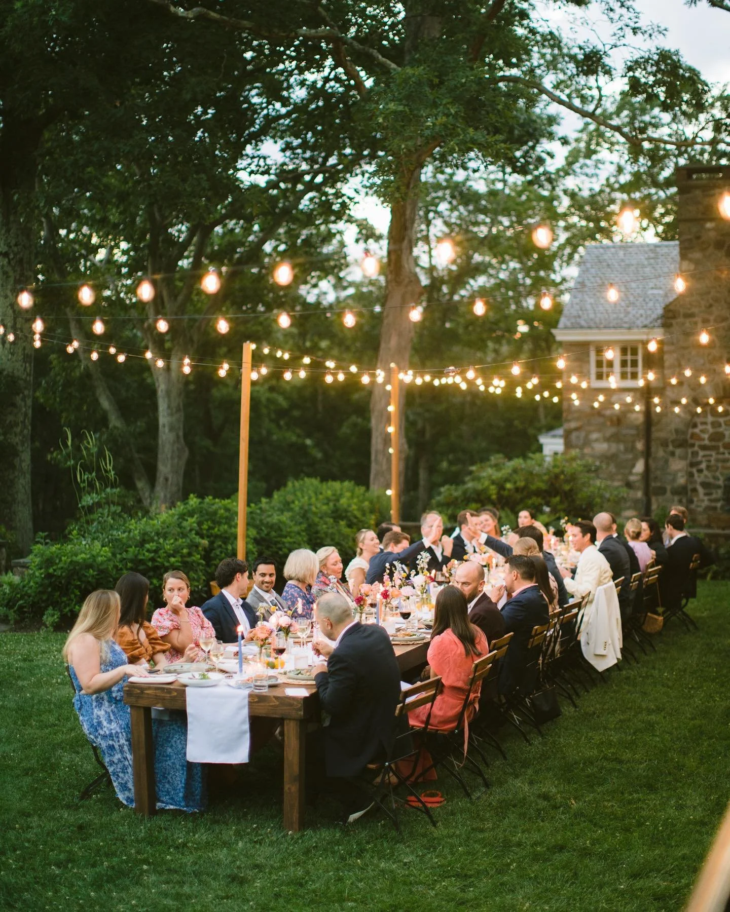 Long outdoor dinner table with guests, lit string lights, trees, and a stone house in the background.
