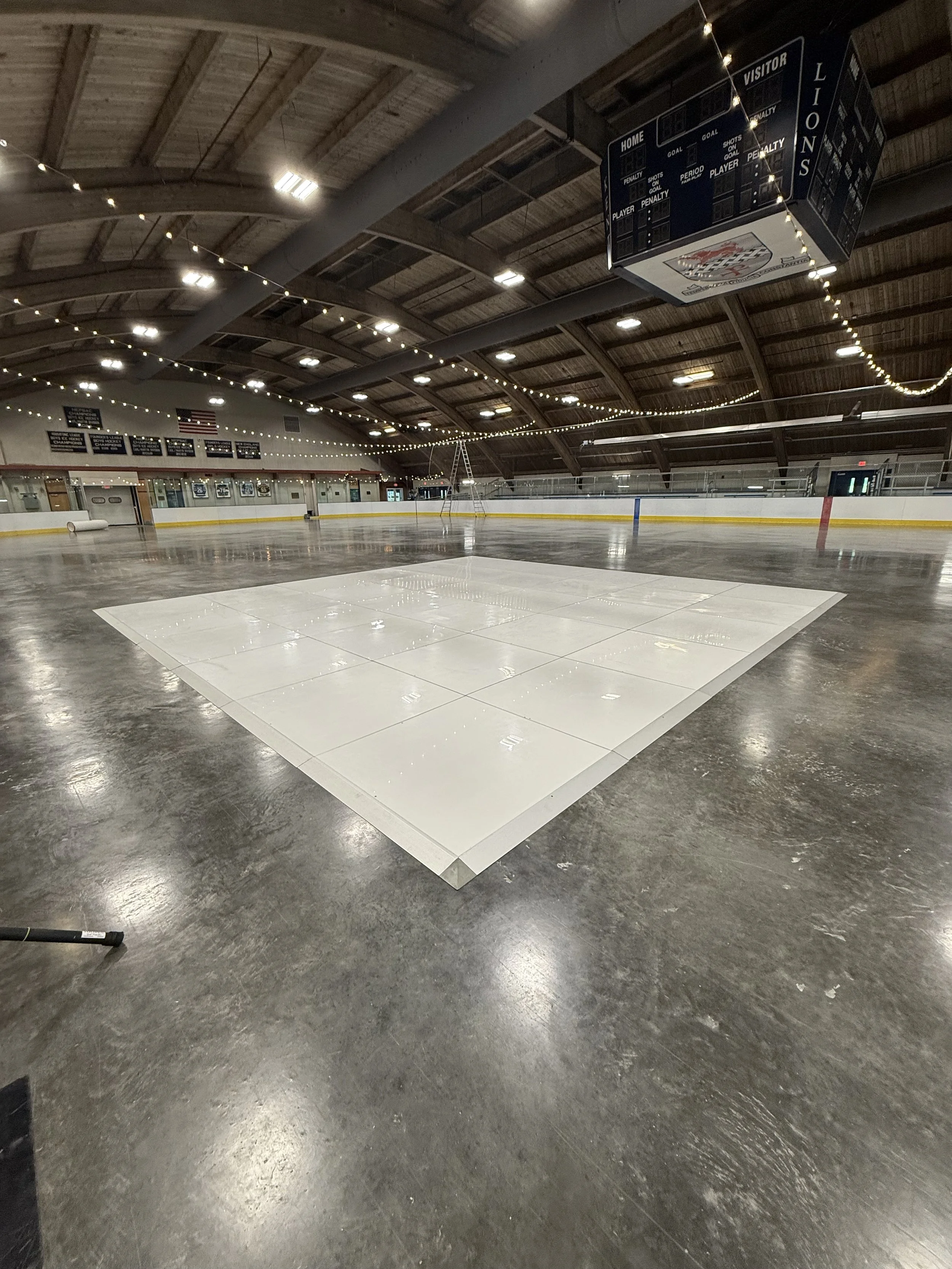 Empty indoor ice skating rink with a white tiled section on the ice surface, string lights hanging from the ceiling, and a scoreboard hanging above the rink.