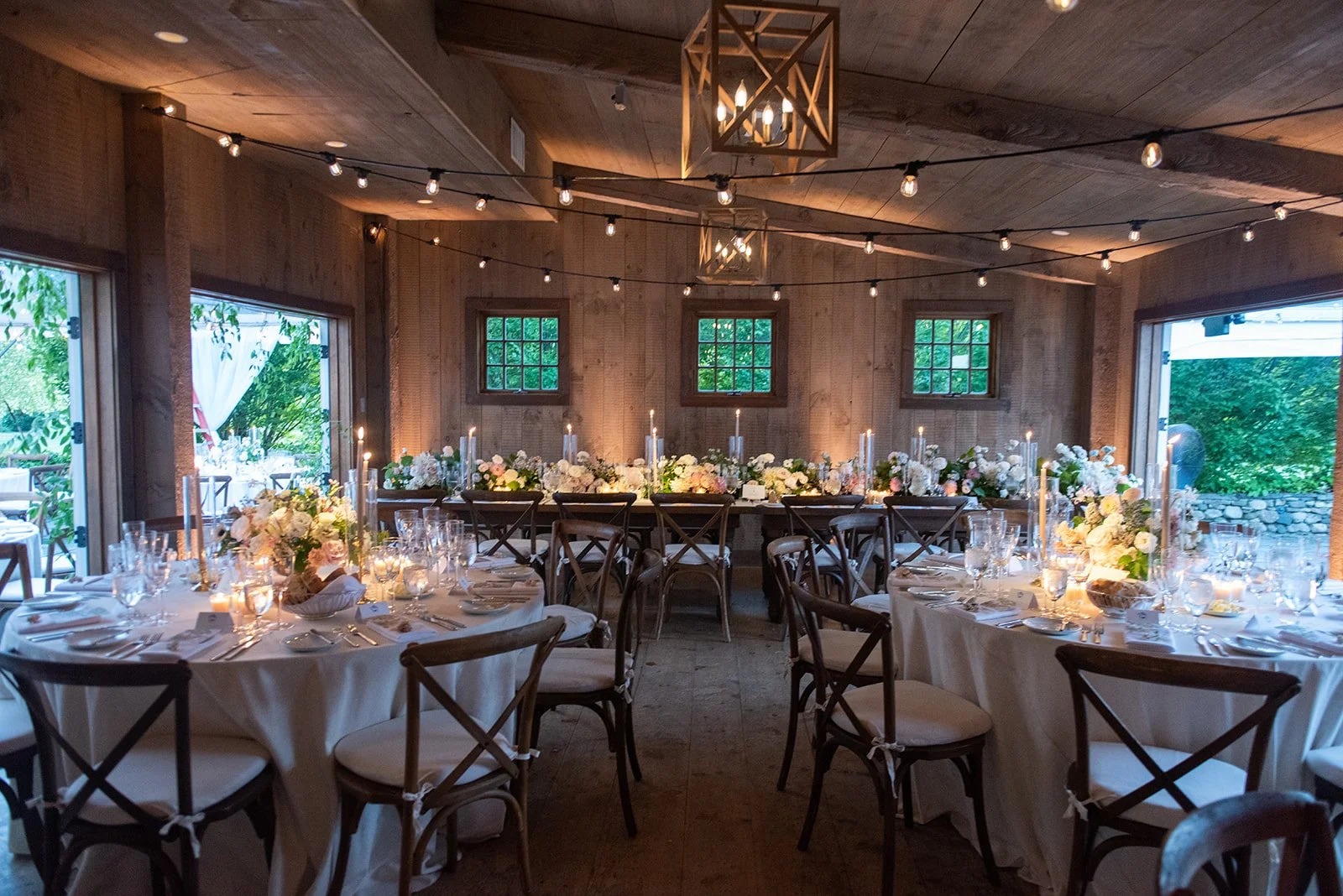 Elegant wedding reception setup inside a wooden venue with tables decorated with white tablecloths, floral centerpieces, candles, and glassware, illuminated by string lights and chandeliers.