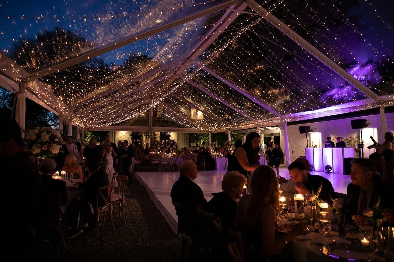 An indoor celebration event with a transparent ceiling decorated with string lights, a space for dancing, and elegantly set tables with candles and floral arrangements.