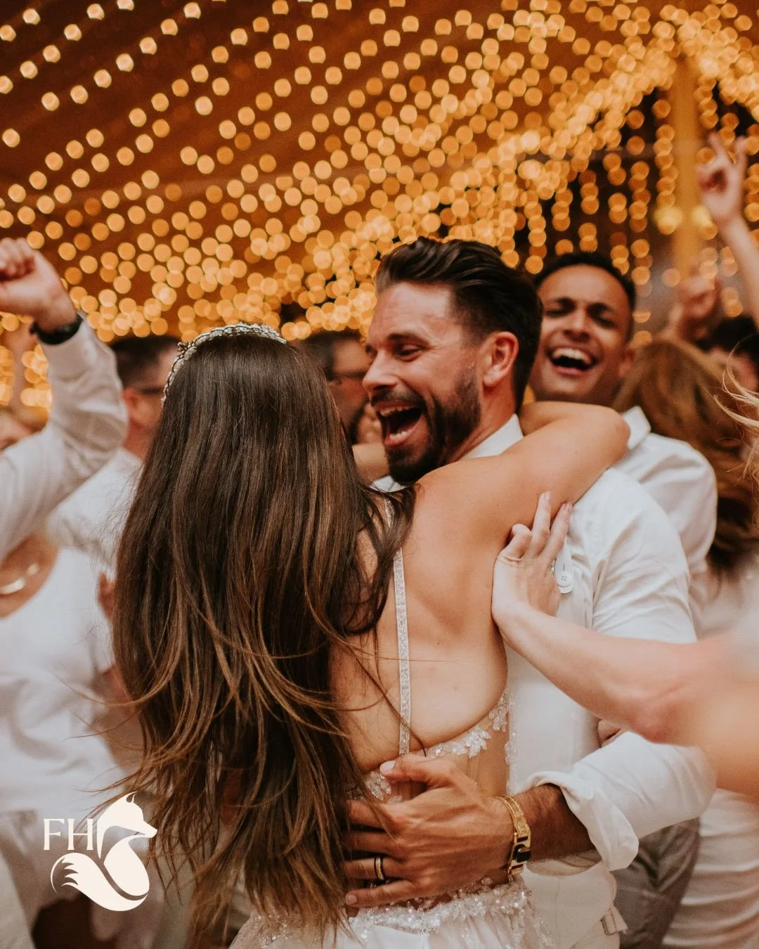 A joyful couple is dancing closely at their wedding reception, surrounded by friends. The background is decorated with warm, string lights.