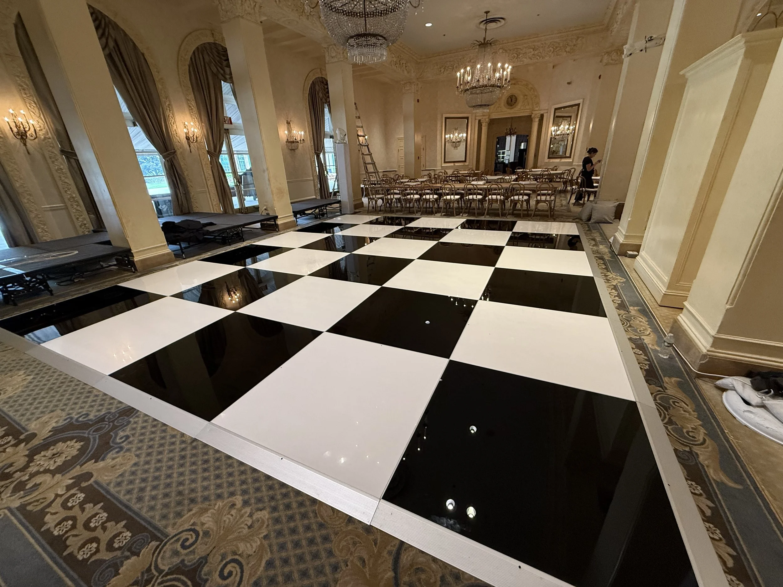 A spacious ballroom with a black and white checkered dance floor, ornate chandeliers, gold-framed mirrors, tall windows with draped curtains, and elegant table and chair arrangements in the background.