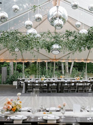 Outdoor event tent decorated with hanging mirror balls and greenery, with a long dining table set with plates, glasses, and floral centerpieces, overlooking a wooded area.