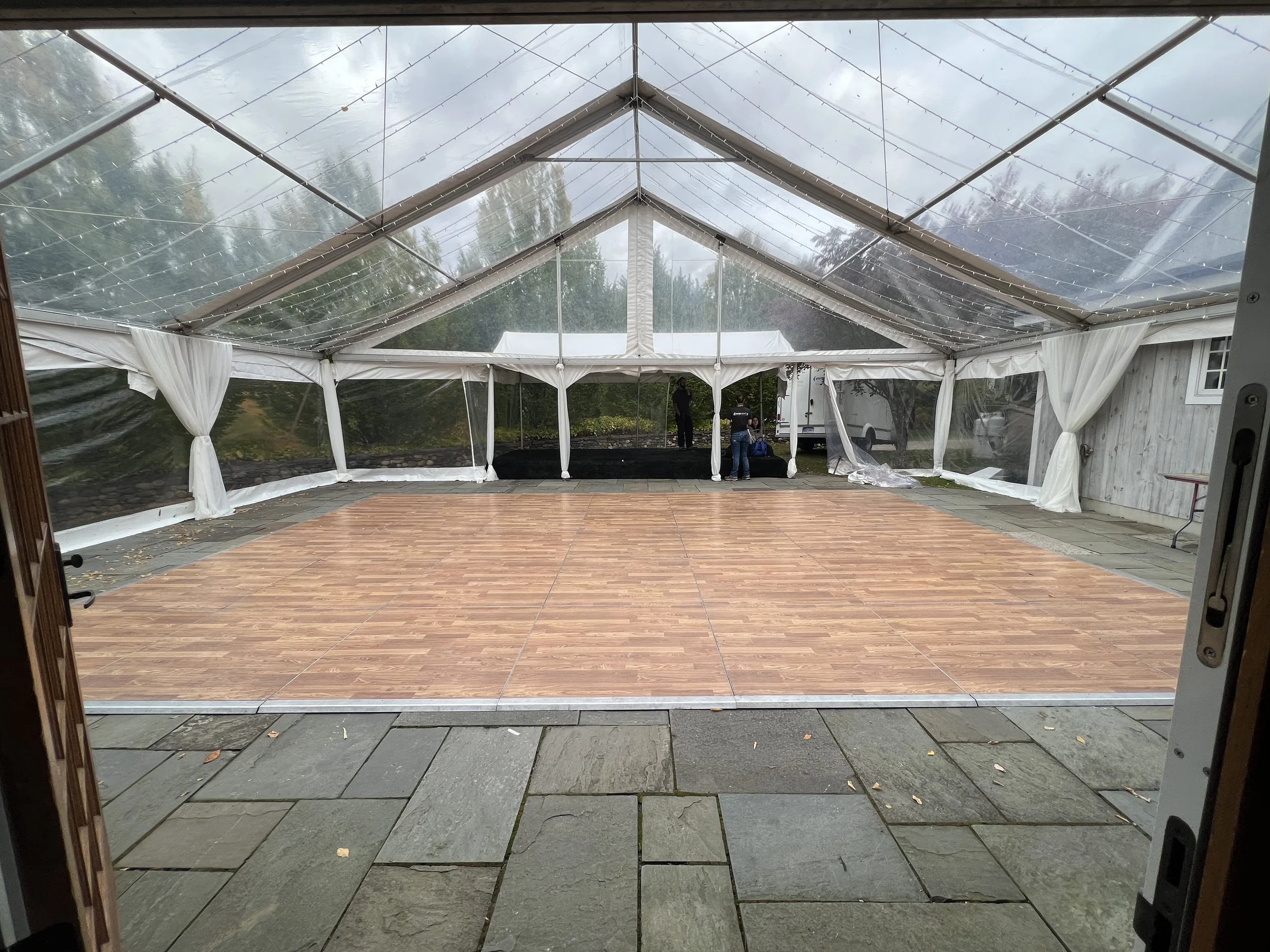 Indoor event space with a transparent tent, a wooden dance floor, and people setting up in the background.