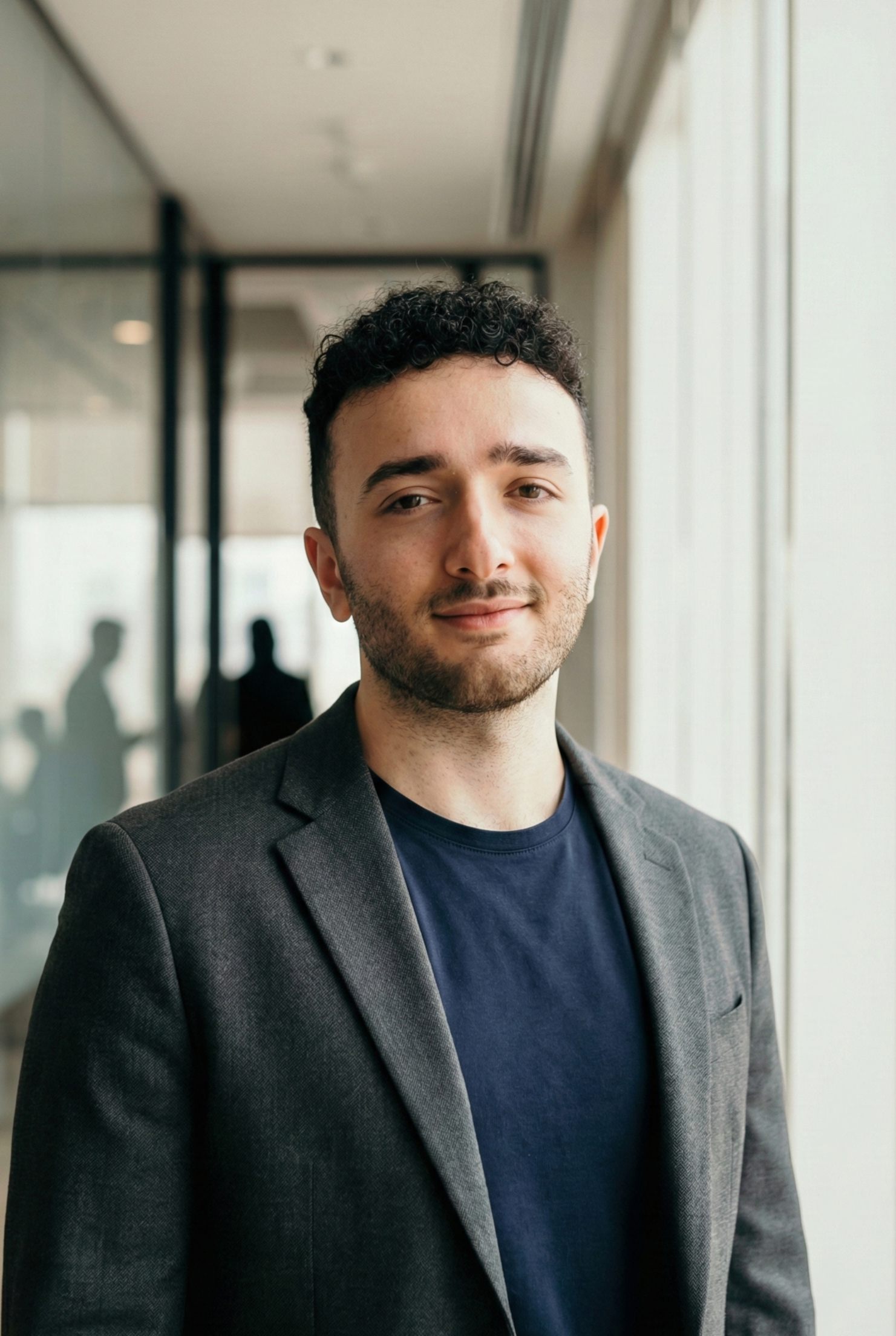 Portrait of a young man with curly hair and light beard, wearing a dark blazer and navy shirt, standing in an office with large windows and blurred reflections of people in the background.