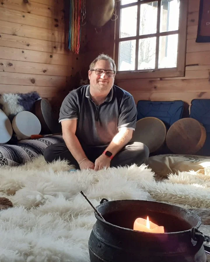 A man sitting inside a cozy wooden room near a candle on a fur rug with pillows and drums in the background.