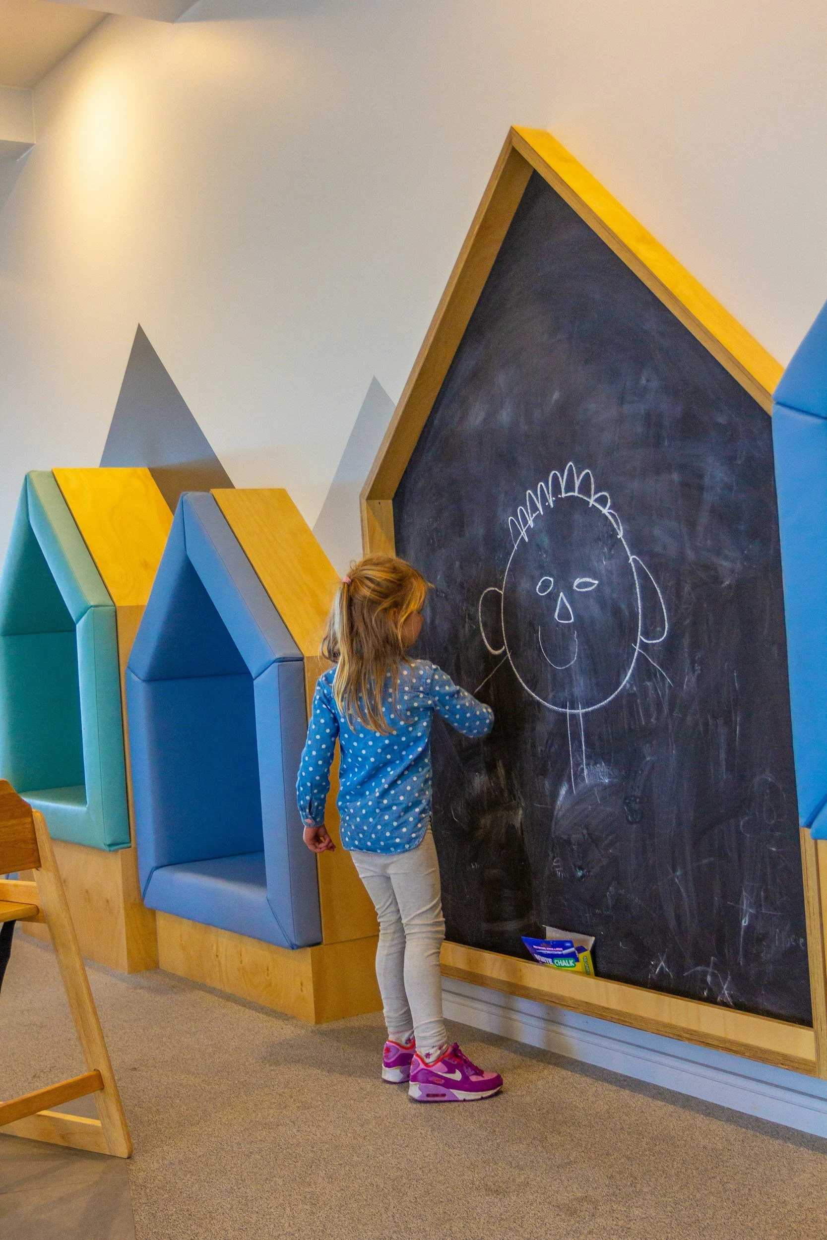 A young girl in a blue polka dot shirt and pink sneakers drawing a face on a large black chalkboard in a colorful indoor play area.