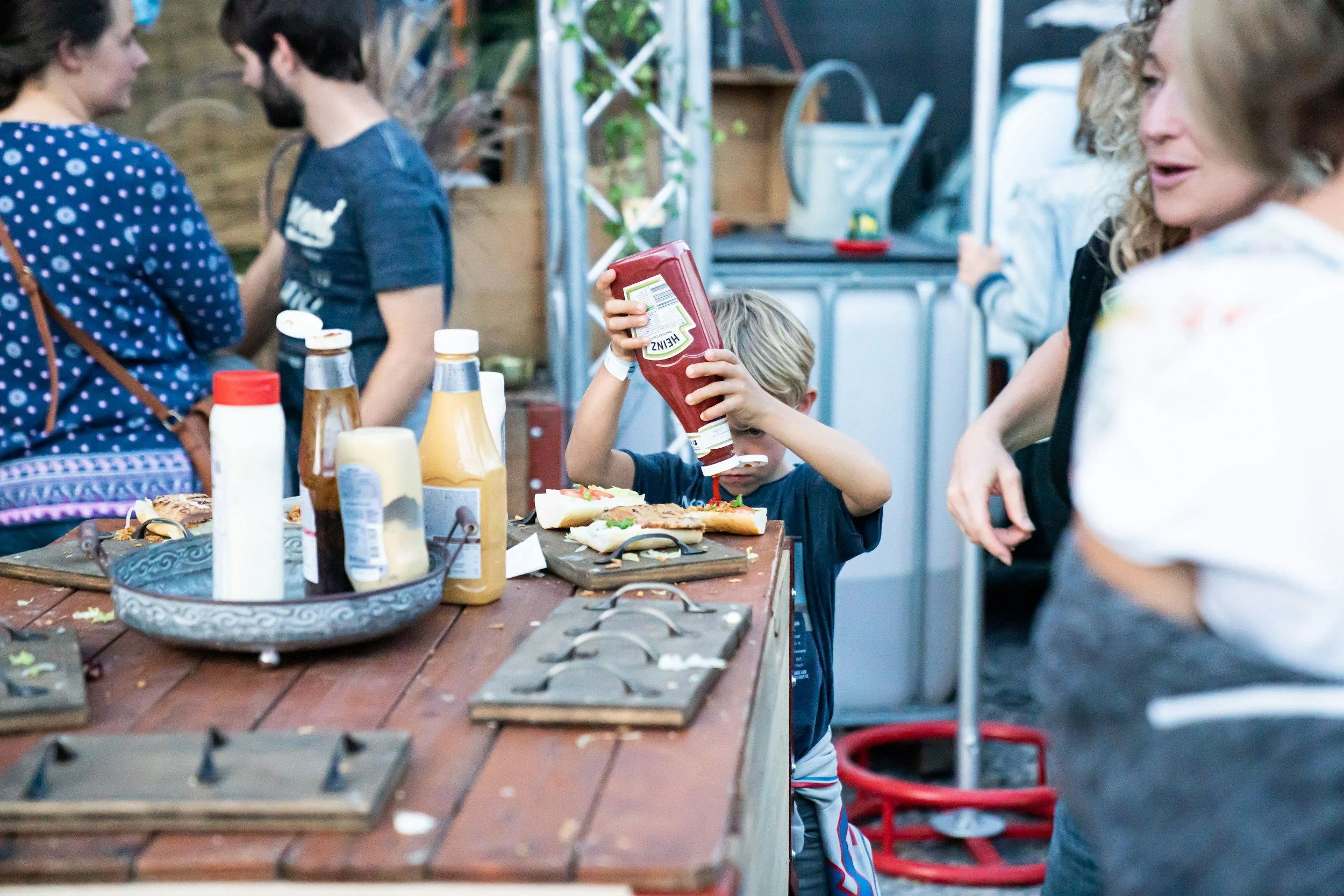 Child pouring ketchup on hot dogs at outdoor gathering with adults and picnic table.