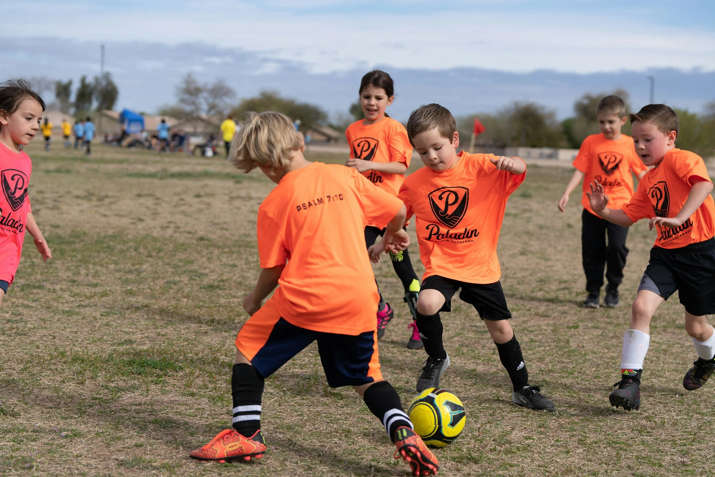 Children playing soccer on a field, wearing orange and pink jerseys with 'Paladin' logos, engaged in a game under a cloudy sky.