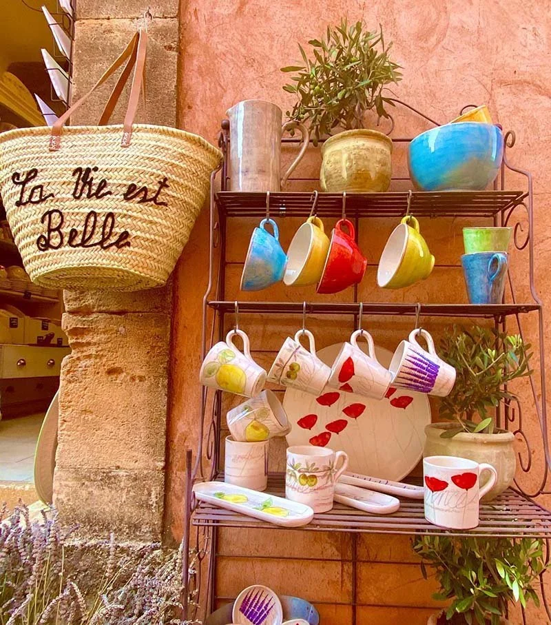 Colorful ceramic mugs and bowls hanging on a metal rack against a pink wall, with potted plants and a woven tote bag nearby.