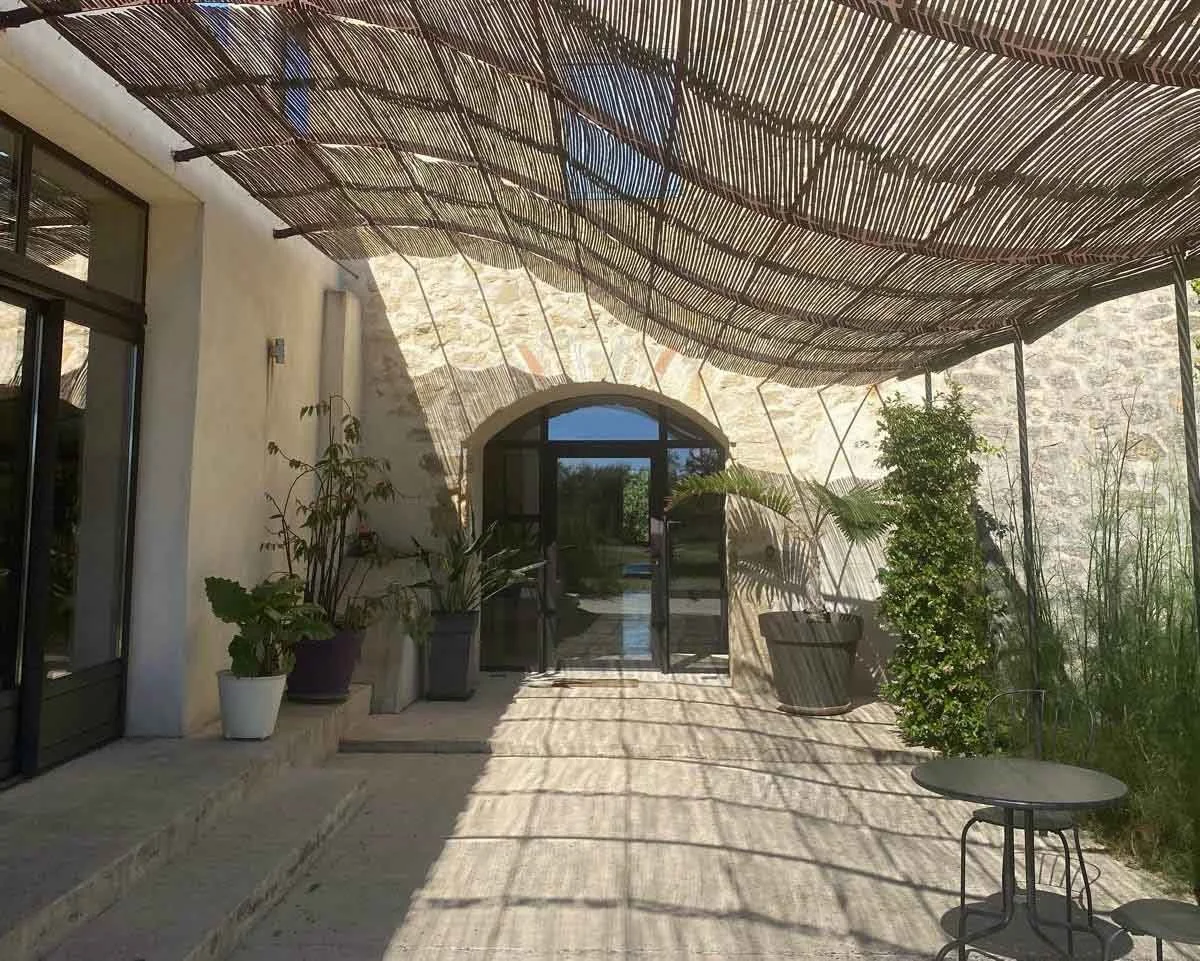 Patio area with potted plants, a small round table with two chairs, and a woven shade structure overhead, casting patterned shadows on the ground. There is a glass door and window on the building's wall.