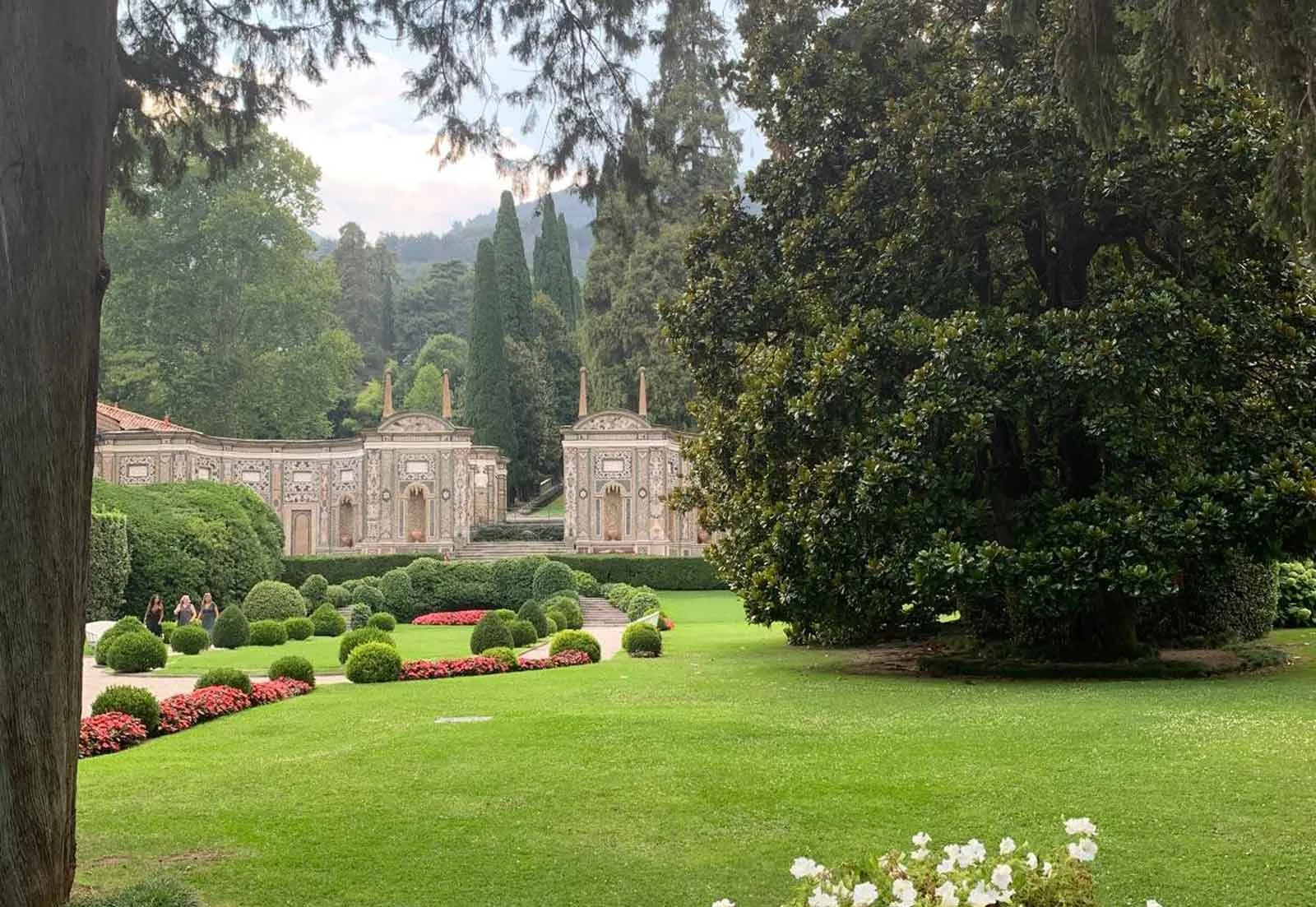 A lush green garden with neatly trimmed bushes and flower beds, and an ornate historic building with decorative architecture in the background, surrounded by tall trees.