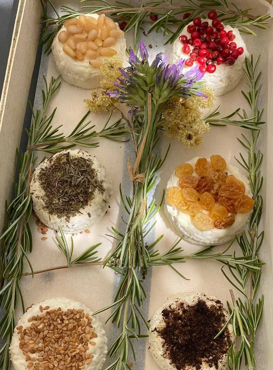 Six small cheese or cracker rounds topped with various toppings, arranged on a tray decorated with sprigs of rosemary and centered on a small bouquet of dried flowers.