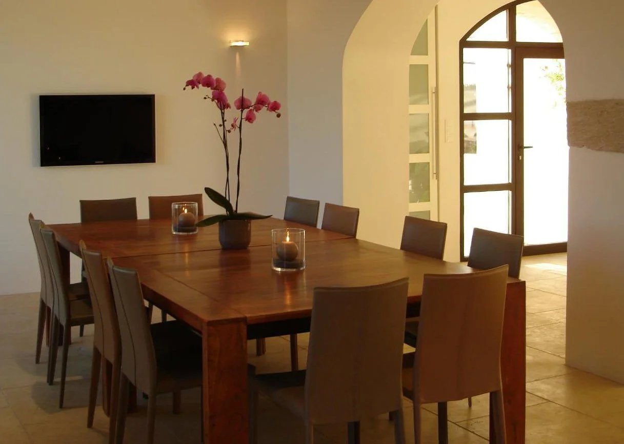Dining room with a wooden table, chairs, a vase with pink orchids, candles, a wall-mounted TV, and an arched glass door letting in sunlight.