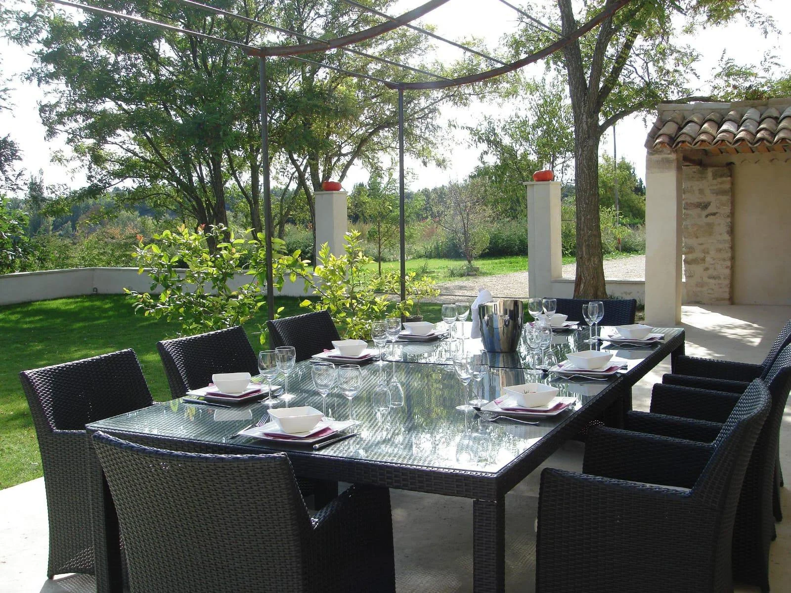 An outdoor patio dining area with a black wicker table set with bowls, glasses, and utensils, surrounded by black chairs, under a shaded area with trees and greenery in the background.