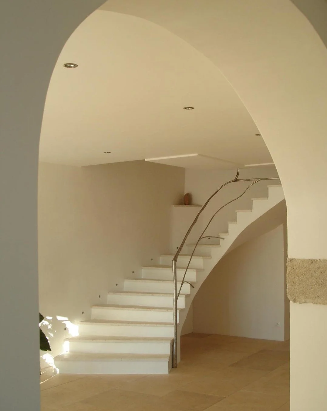 Interior view of a modern staircase with white steps, a curved metal handrail, and an arched doorway leading to a beige room. Recessed ceiling lights and a small decorative object on a shelf are visible.