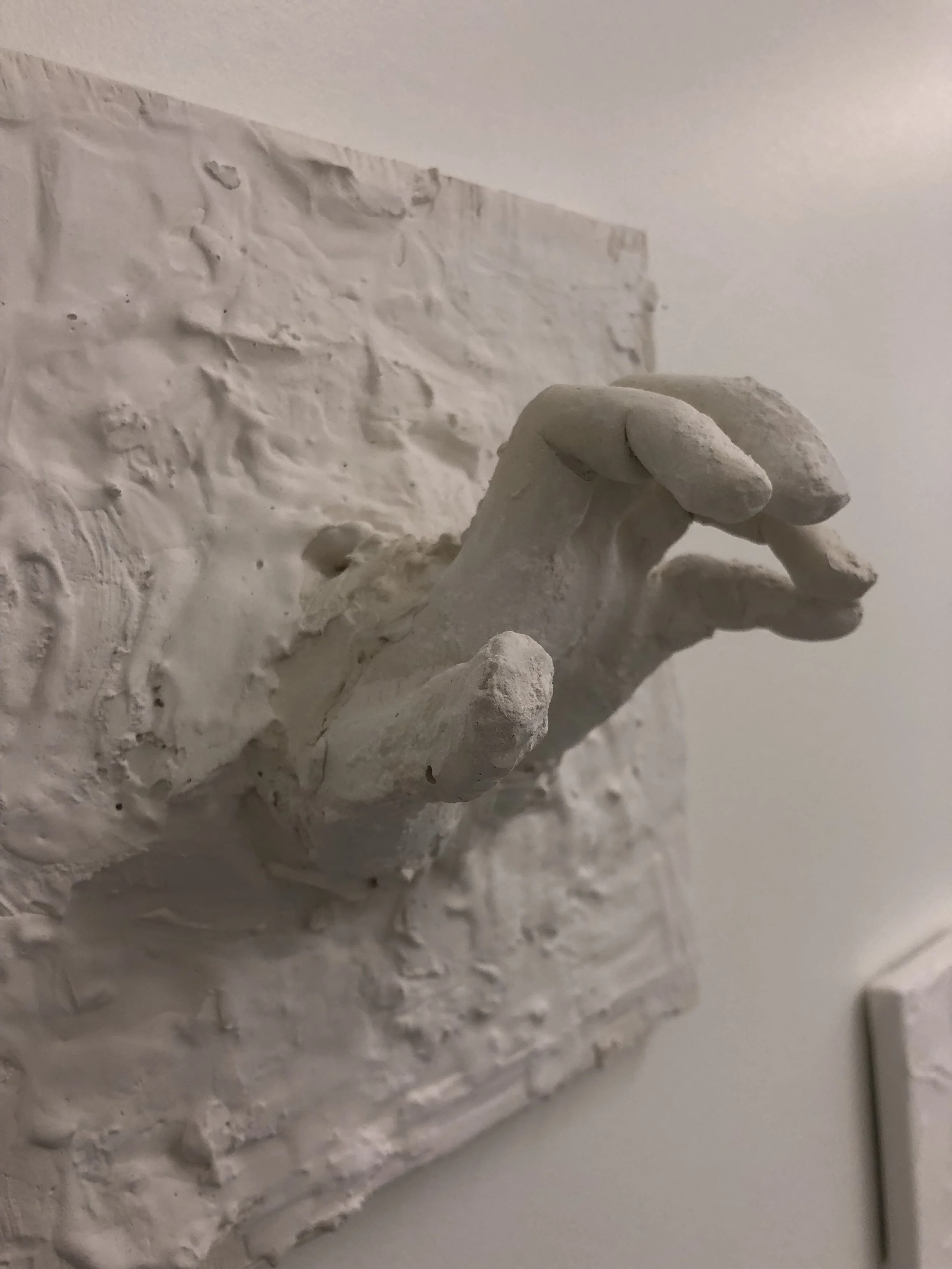 White sculpture of a hand with fingers curled, mounted on a textured white wall.