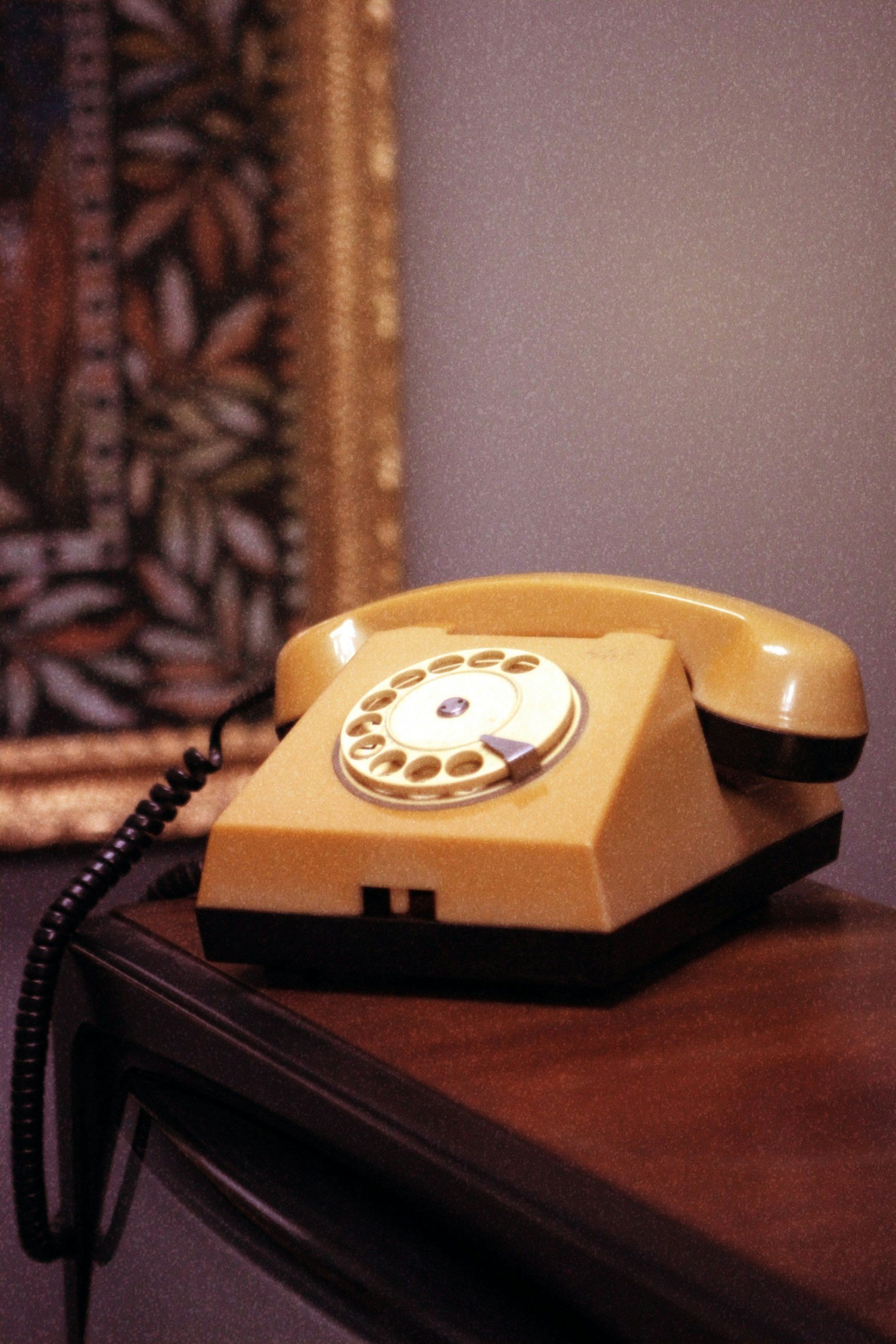 A yellow rotary telephone sitting on a wooden table, with a dark-colored cord attached to it. The background includes a decorative framed artwork.