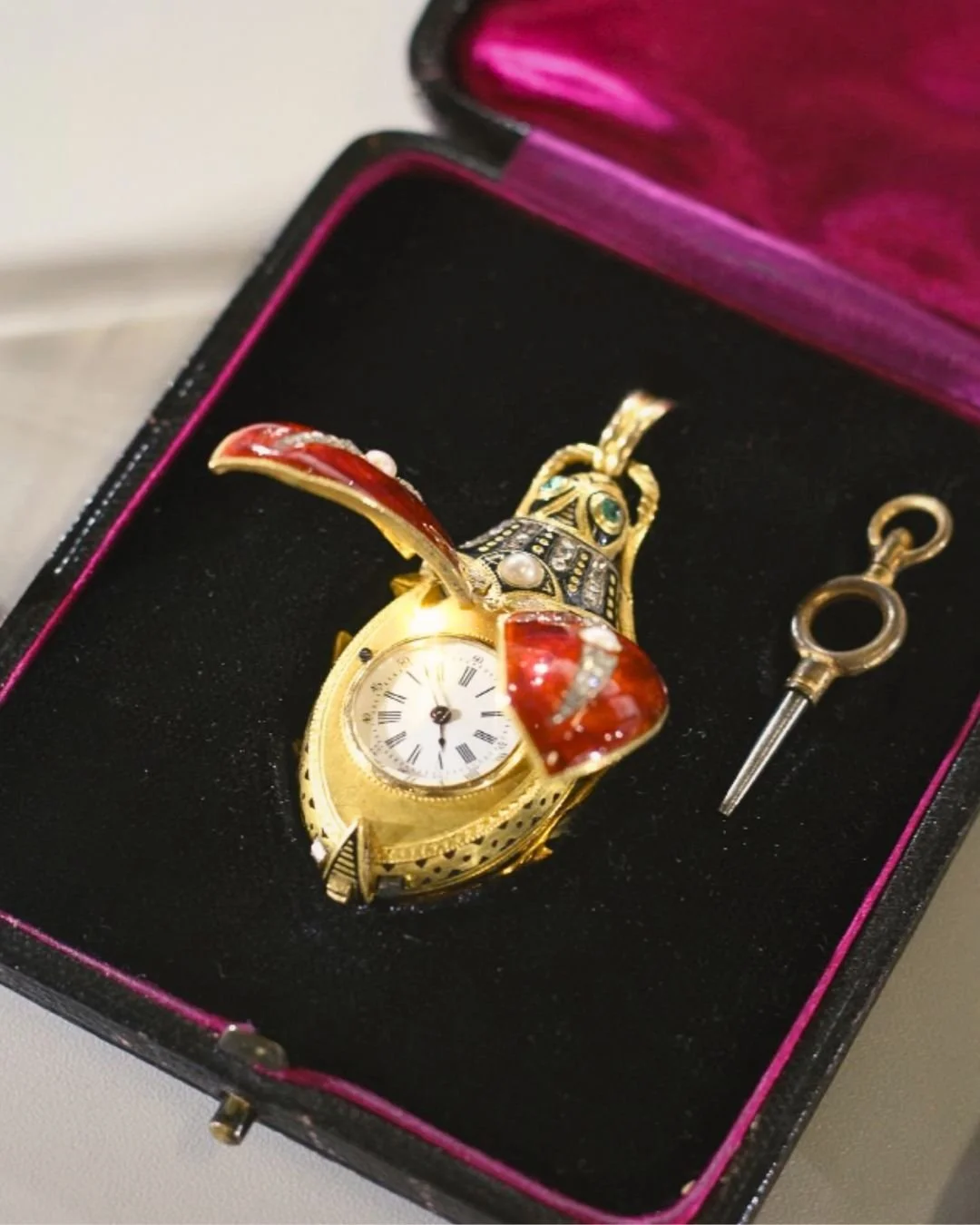 A gold rabbit-shaped watch with a face displaying Roman numerals, decorated with red and pearl accents, inside a jewelry box with a black interior and pink edges, along with a small key or tool.