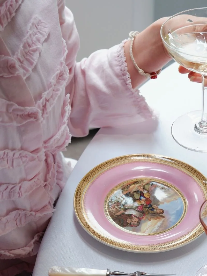 Person wearing a pink frilly blouse holding a glass of wine, with a decorative pink and gold plate on the table.
