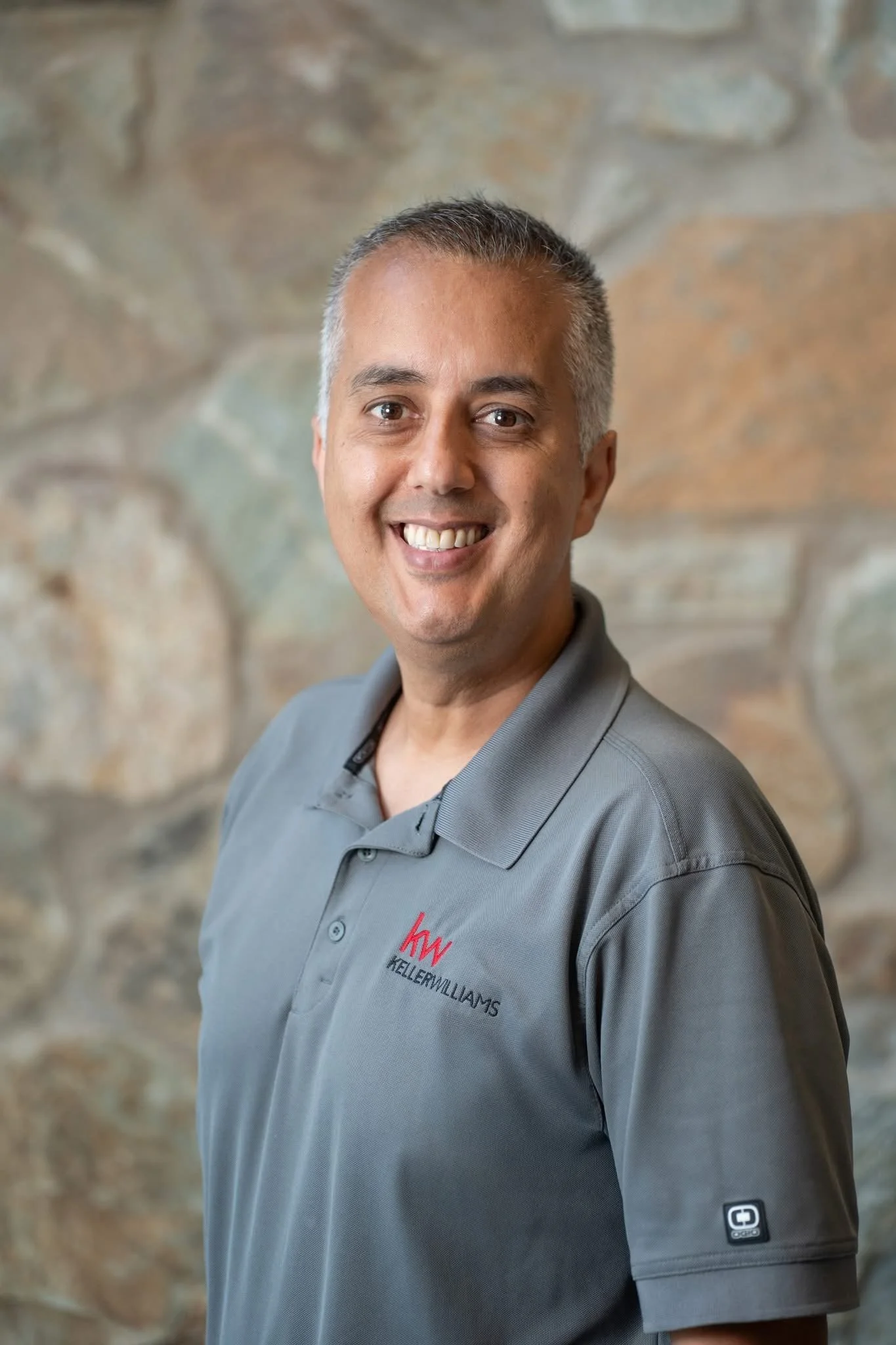 A smiling man wearing a gray polo shirt with a Keller Williams logo, standing in front of a stone wall background.