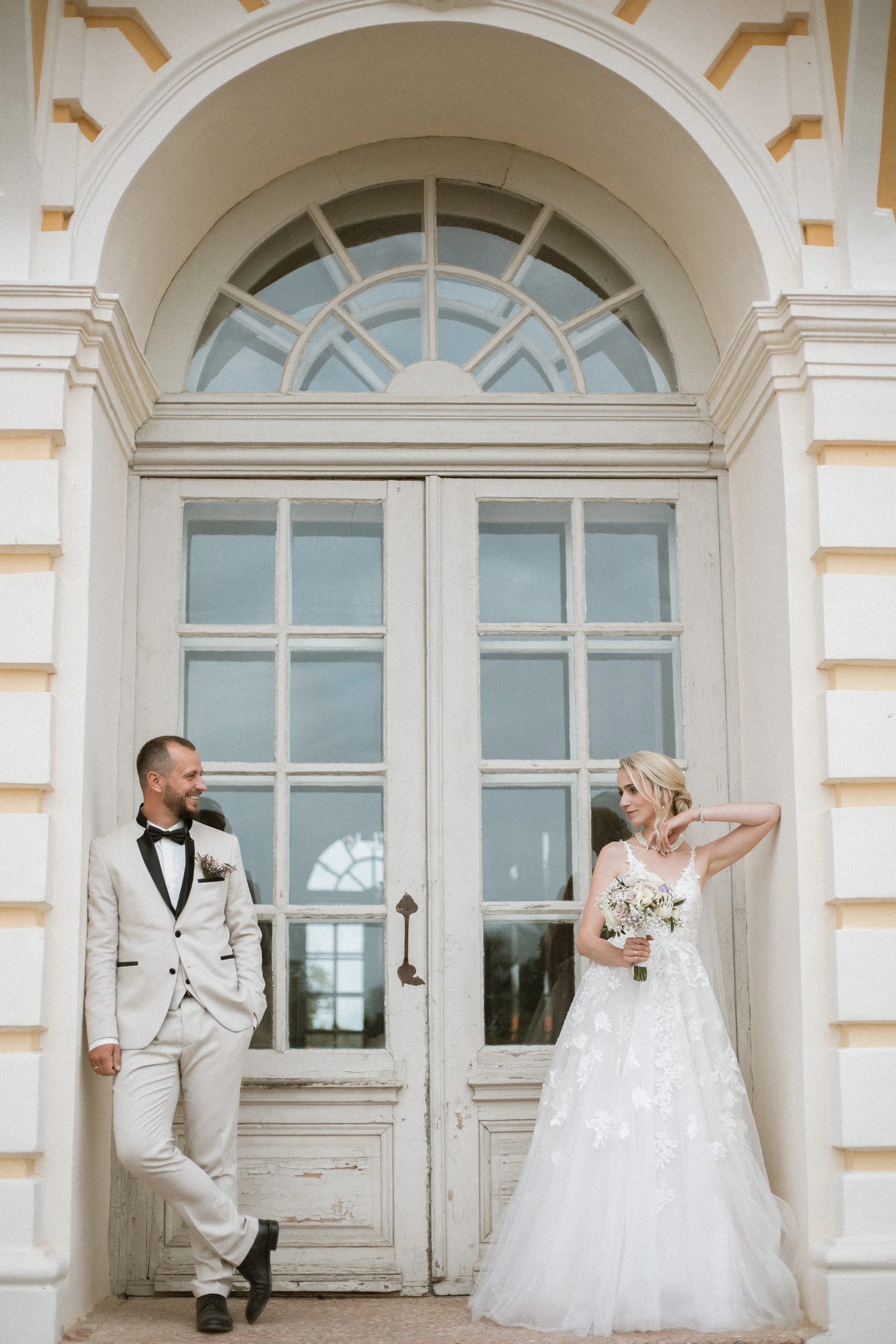A groom in a white tuxedo with a black bow tie and a bride in a white wedding dress holding a bouquet, standing in front of a large, old-fashioned door with glass panes, outside a building.
