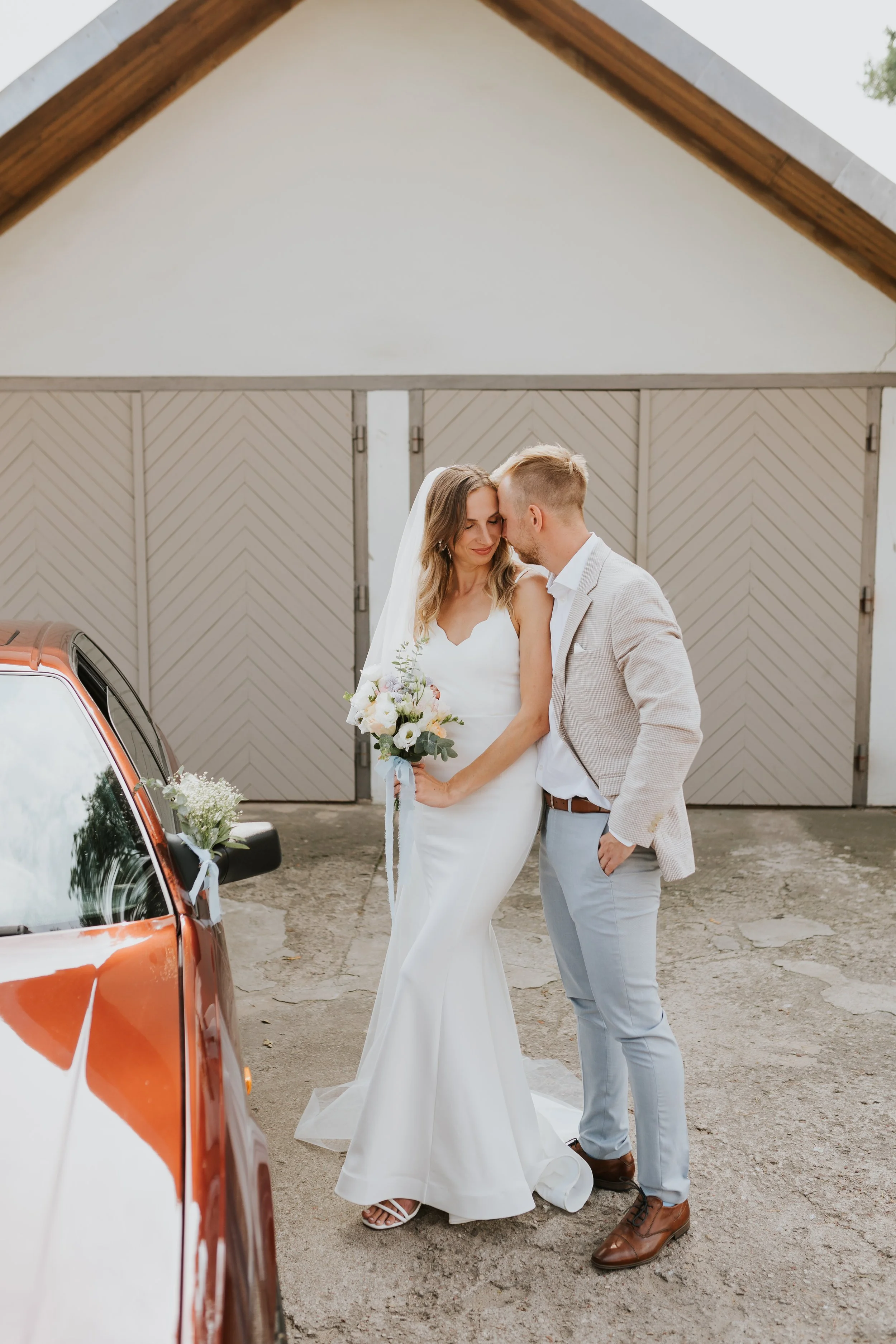 A bride and groom share a tender moment outdoors in front of a garage door, with the bride holding a bouquet of flowers and both dressed in wedding attire.