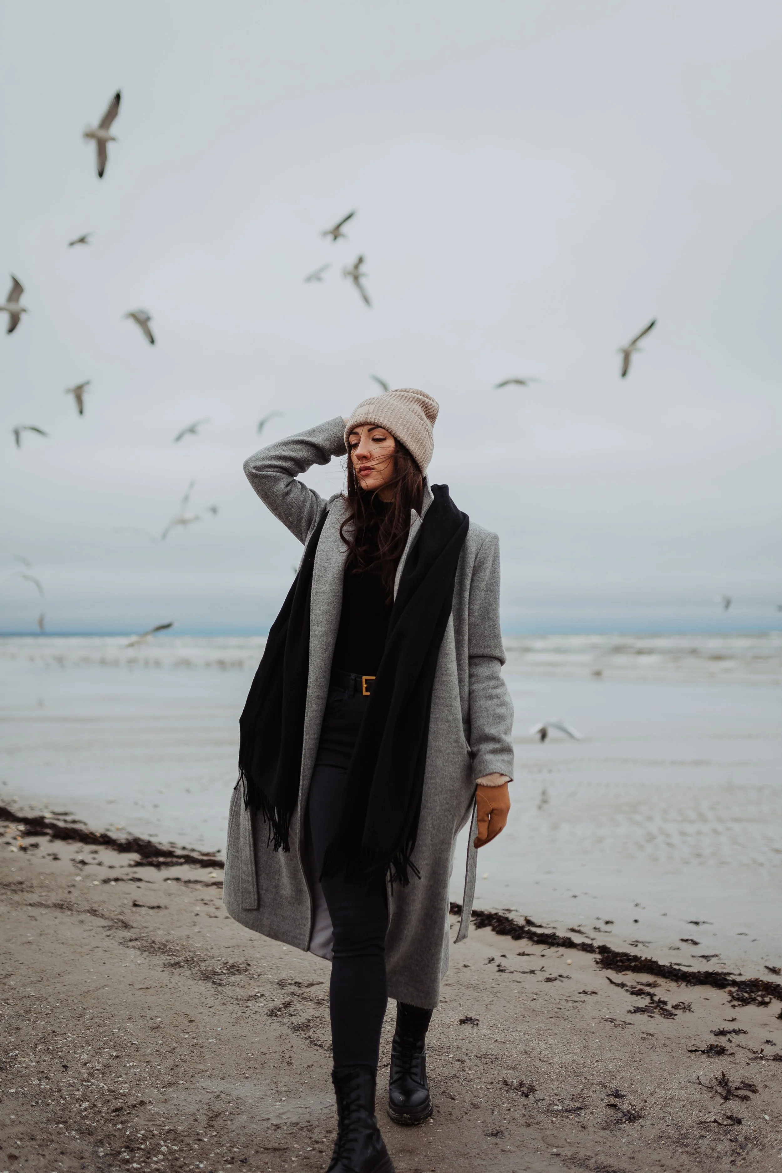 Woman walking on a beach with seagulls flying overhead, dressed warmly in a gray coat, beige beanie, black scarf, black pants, and black boots.