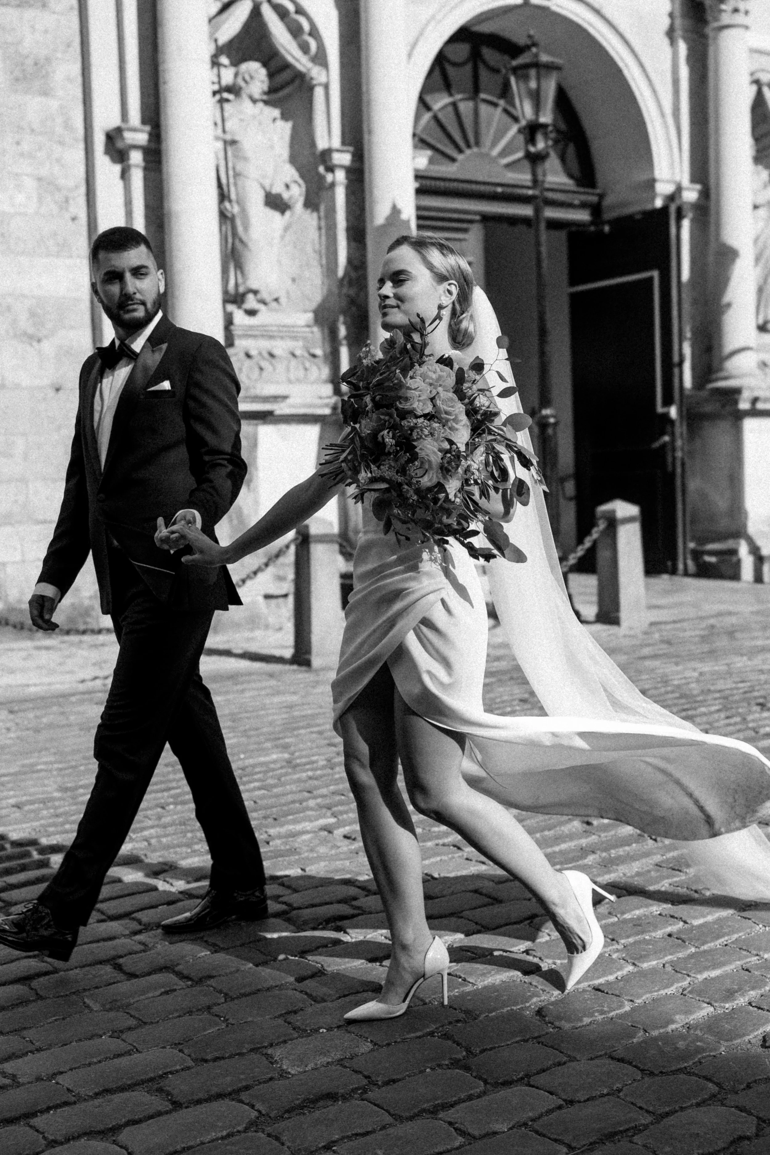 Black and white photo of a bride and groom walking outside, holding hands, with the bride in a strapless wedding dress and high heels, carrying a large bouquet, and the groom in a tuxedo, in front of a historic building with statues and an open door.