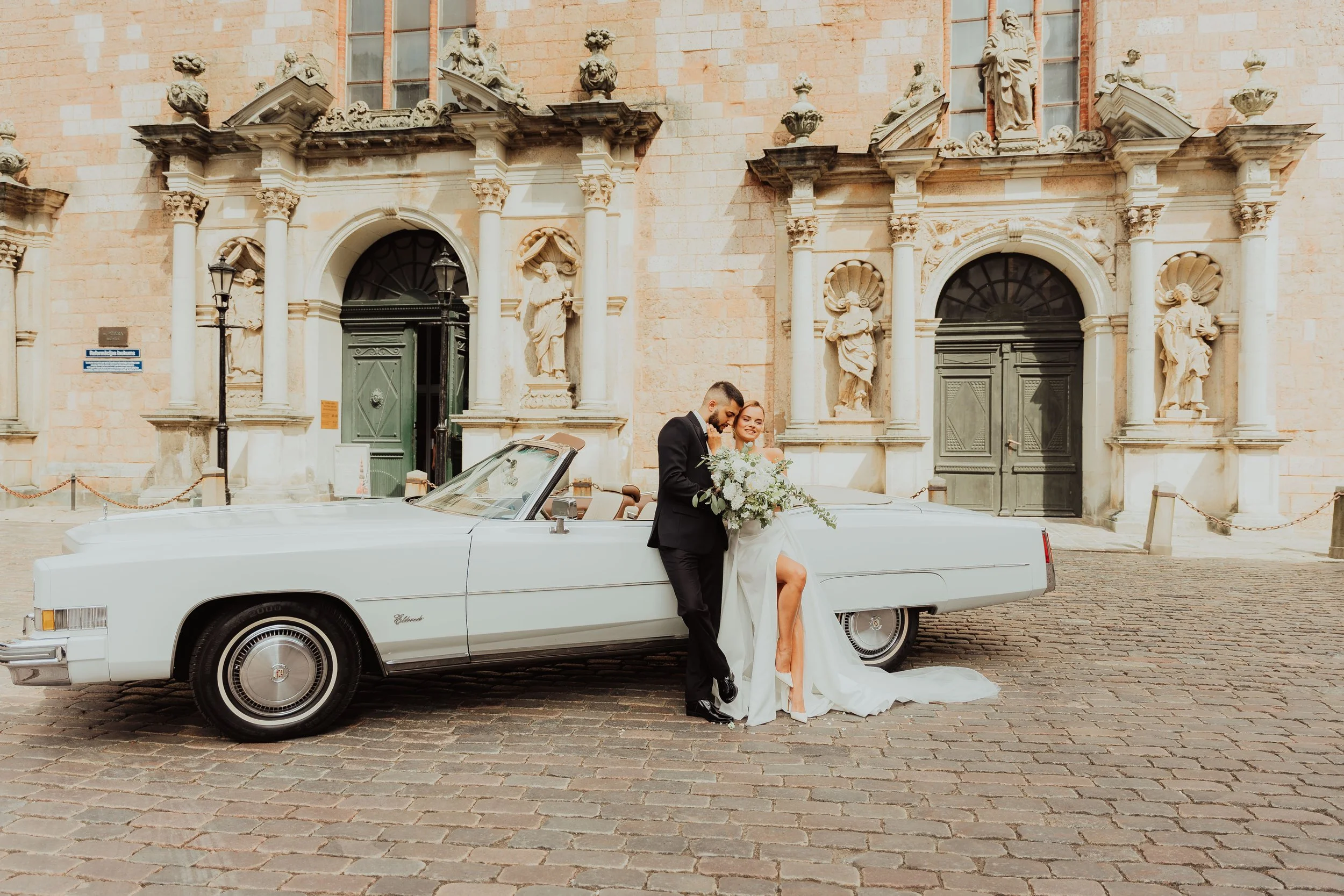 Wedding couple standing next to a white vintage convertible car in front of an ornate historic building with statues and detailed architecture.